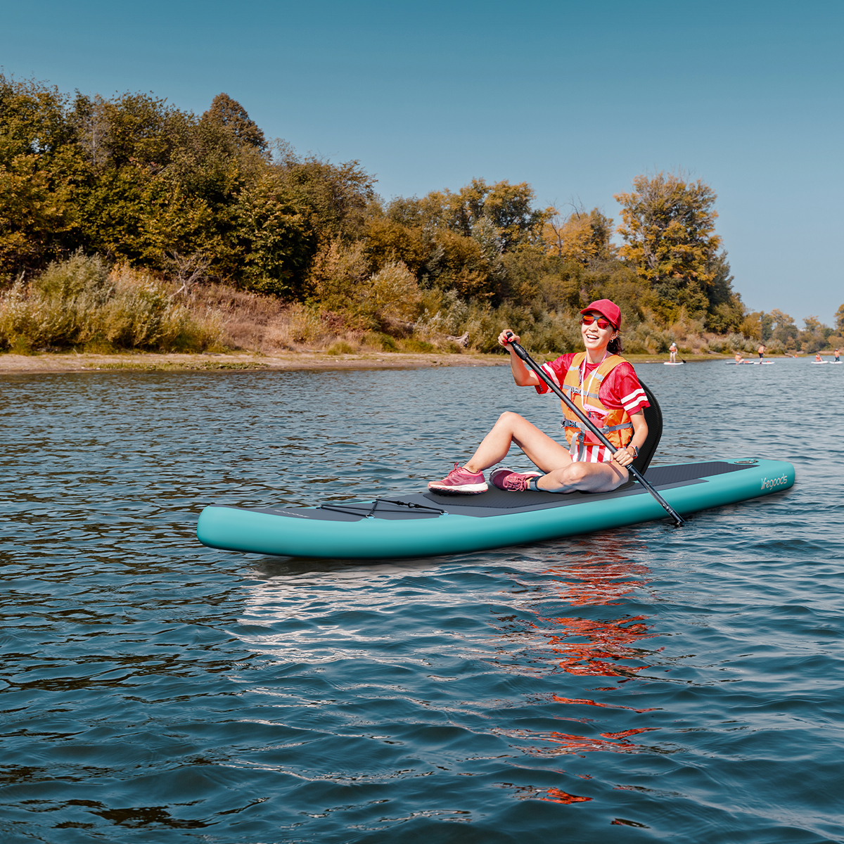 Een vrouw peddelt op een blauw stand-up paddleboard op een rivier met bomen op de achtergrond.