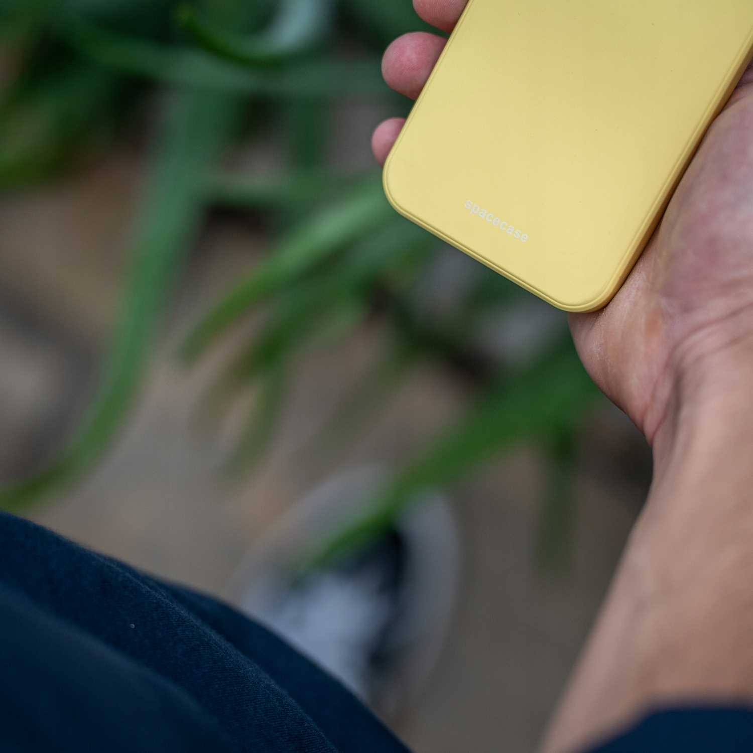 Osoba trzyma żółte etui na telefon z napisem „spacecase”, w tle zielone rośliny.