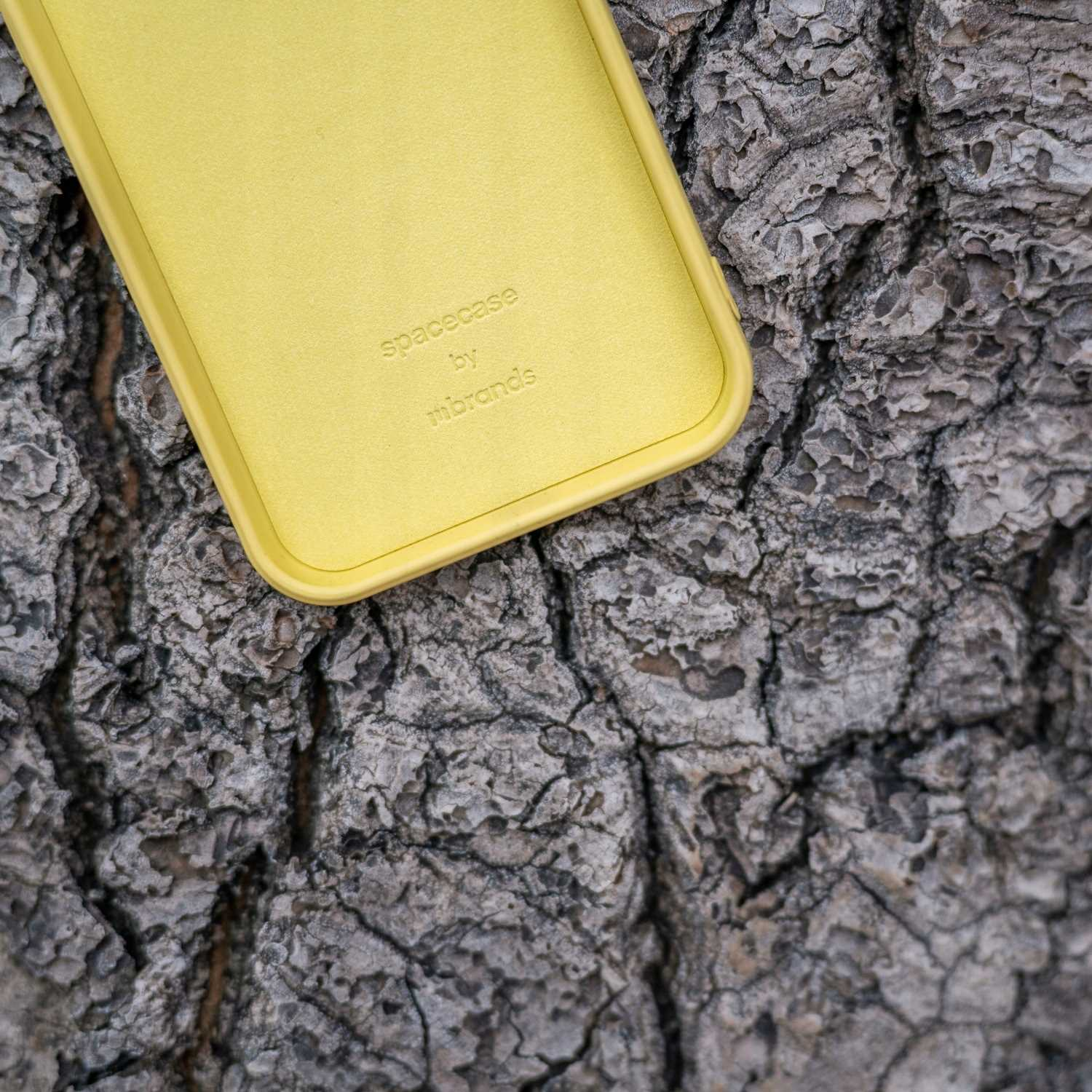 Żółte etui na telefon z napisem „spacecase” znajduje się na szorstkim, szarym pniu drzewa.