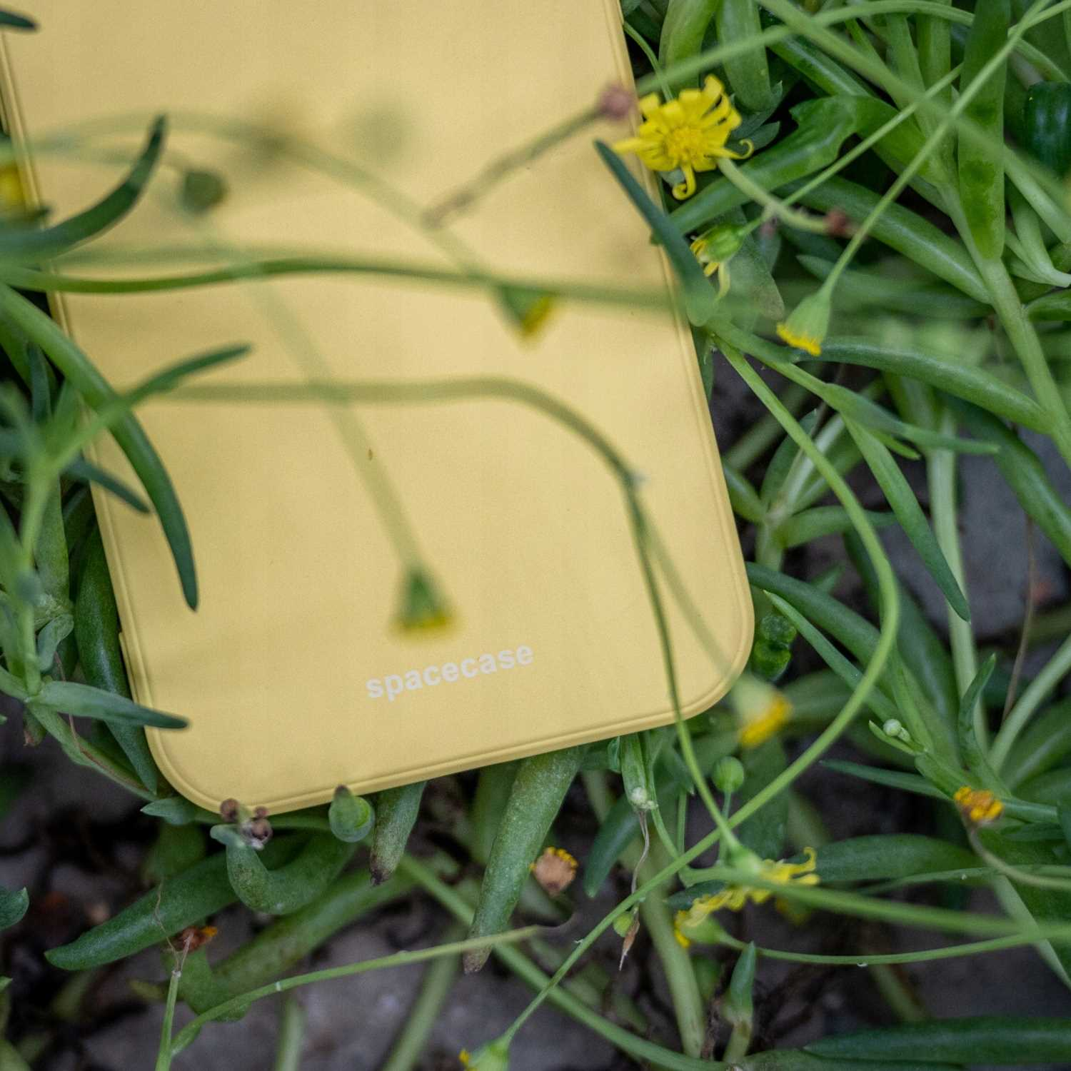Żółte etui na telefon z napisem „spacecase” znajduje się w zielonych roślinach z małymi żółtymi kwiatami.