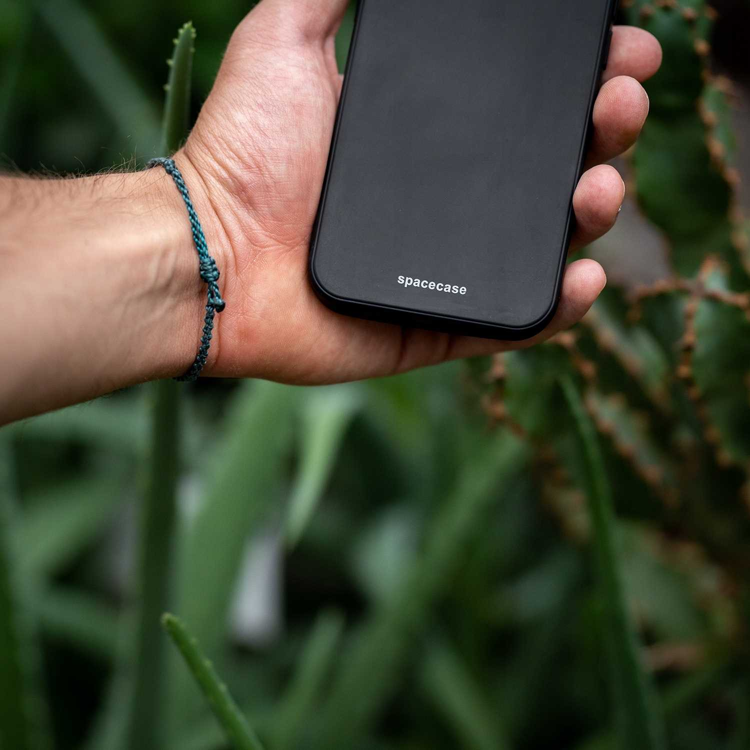 Dłoń trzymająca czarny telefon z 'spacecase', turkusowa bransoletka na nadgarstku, zielone tło.