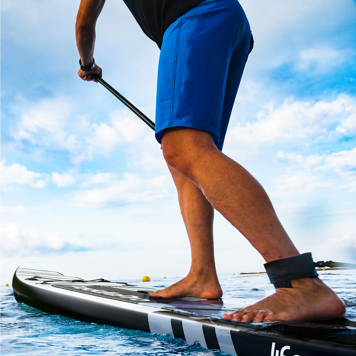 Une personne se tient sur une planche de paddle dans l'eau, portant un short bleu et une sangle de cheville, tenant une pagaie.