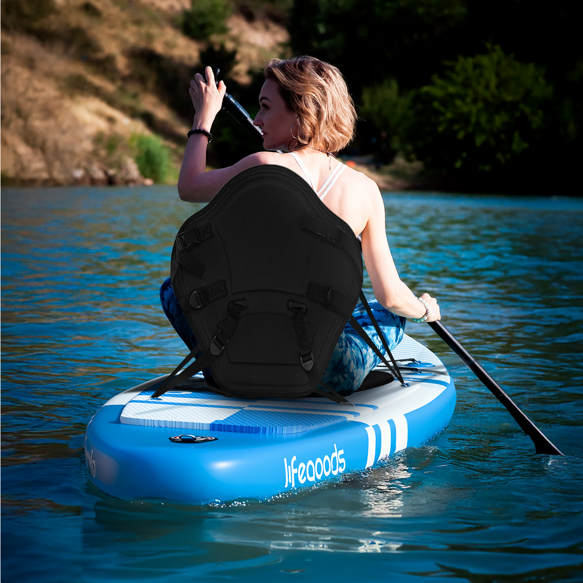 Frau sitzt auf einem blau-weißen Paddleboard mit einem schwarzen Sitz. Sie hält ein Paddel.