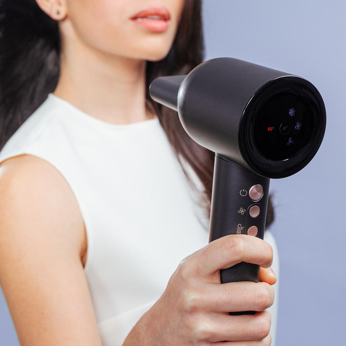 Een vrouw die een zwarte haardroger vasthoudt. Ze kijkt naar de droger, op een blauwe achtergrond.