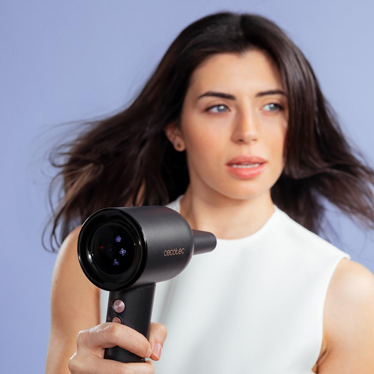 Een vrouw die een zwarte haardroger vasthoudt en naar rechts kijkt. Blauwe achtergrond. De droger heeft een display.