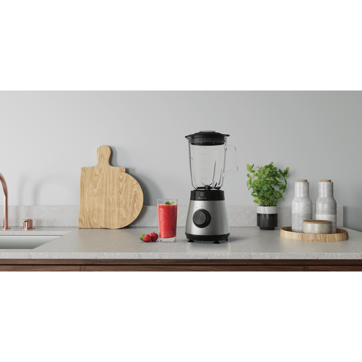 Batidora plateada Electrolux sobre una encimera con un batido rojo, una tabla de cortar y dos botellas.