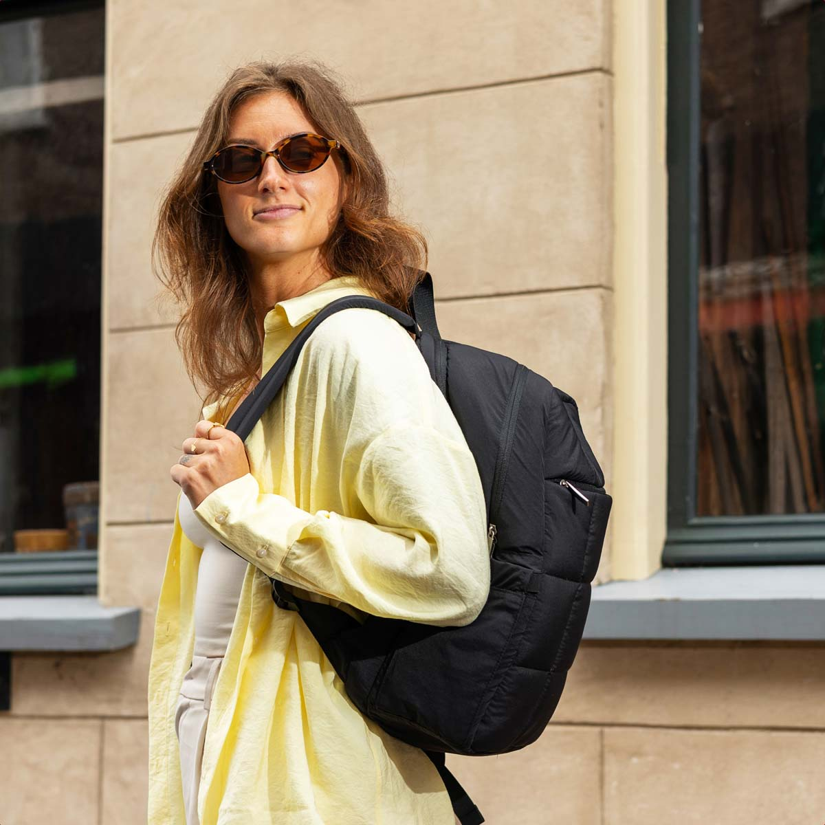 A woman wearing sunglasses and a yellow shirt is wearing a black quilted backpack.