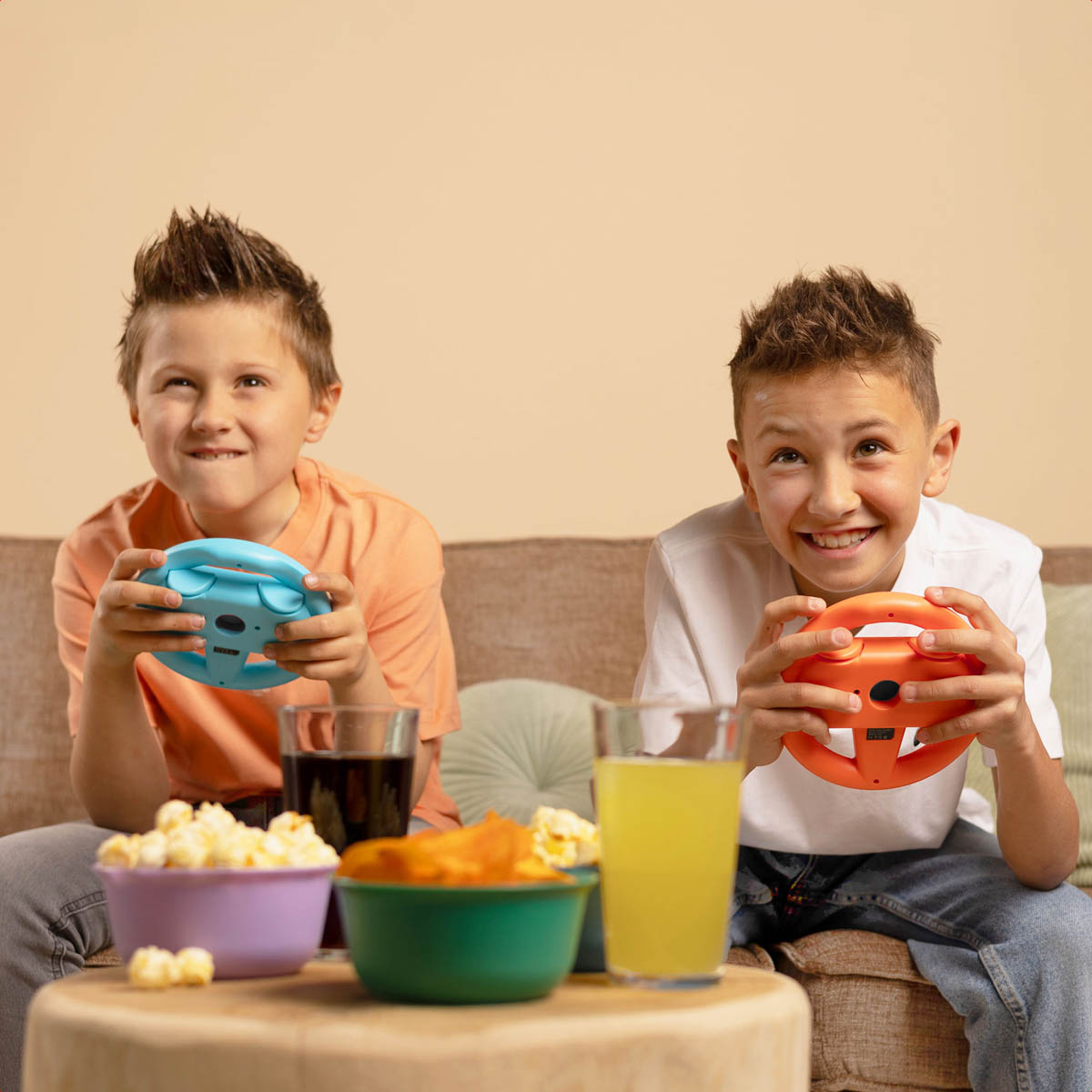 Due ragazzi stanno giocando ai videogiochi con accessori per volante blu e arancioni, con snack e bevande su un tavolo.