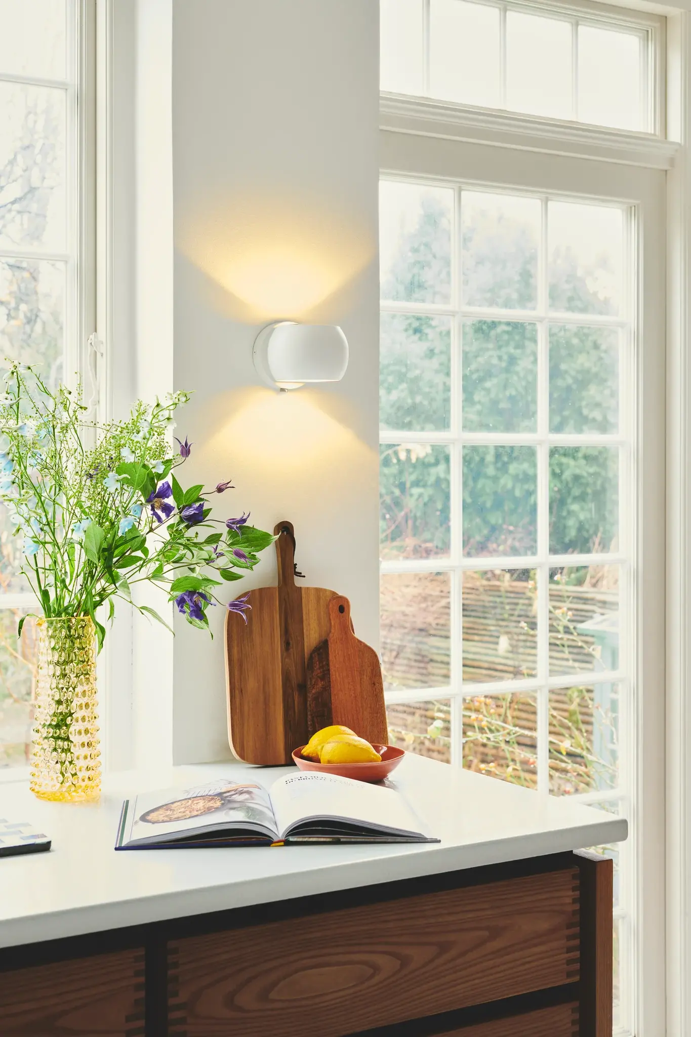 Küchenszene mit Wandleuchte, Blumenvase, Zitronen und einem Kochbuch auf einer Theke.