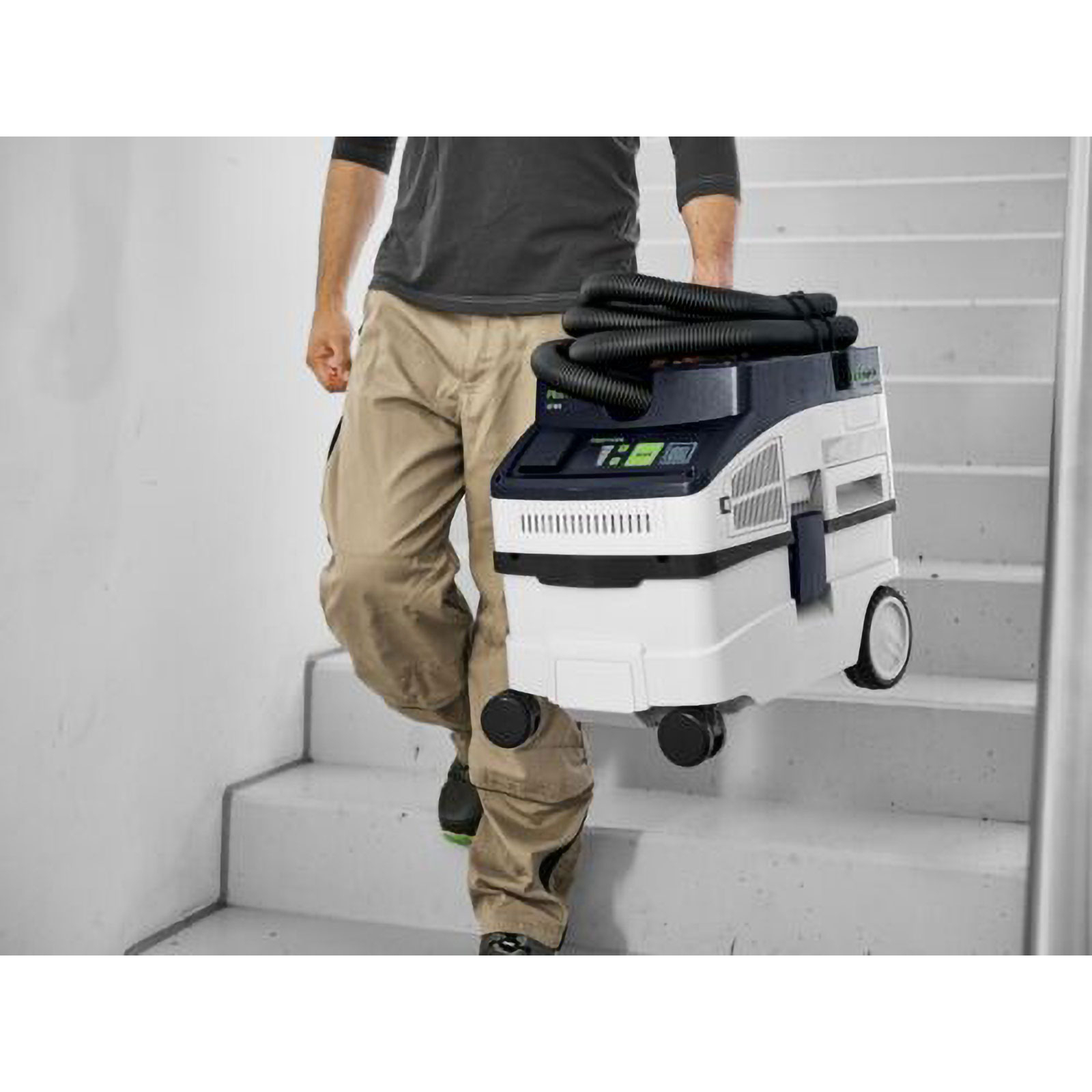 Uomo che trasporta un aspirapolvere Festool bianco e blu scuro sulle scale. L'aspirapolvere ha tubi neri.