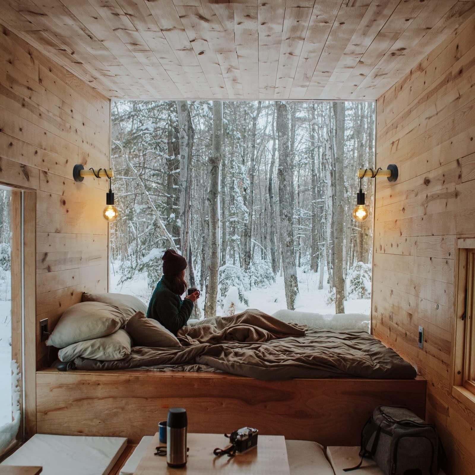 Scena interna di una cabina con una persona a letto vicino a una grande finestra che si affaccia su una foresta innevata. Sono montate due lampade da parete.