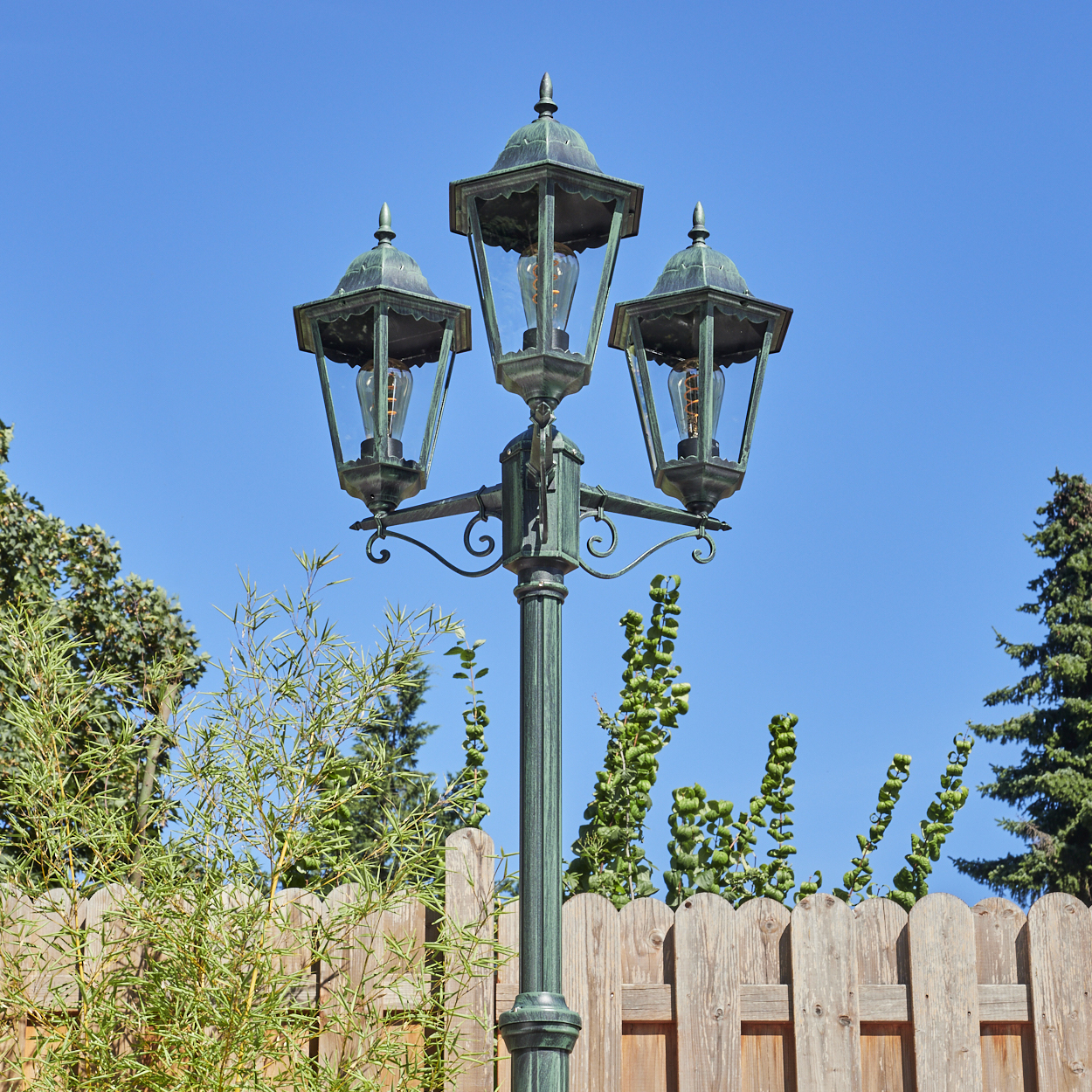 Eine Straßenlaterne im Vintage-Stil mit drei Lampen steht in einem Garten mit einem Holzzaun und Grün unter einem klaren blauen Himmel.