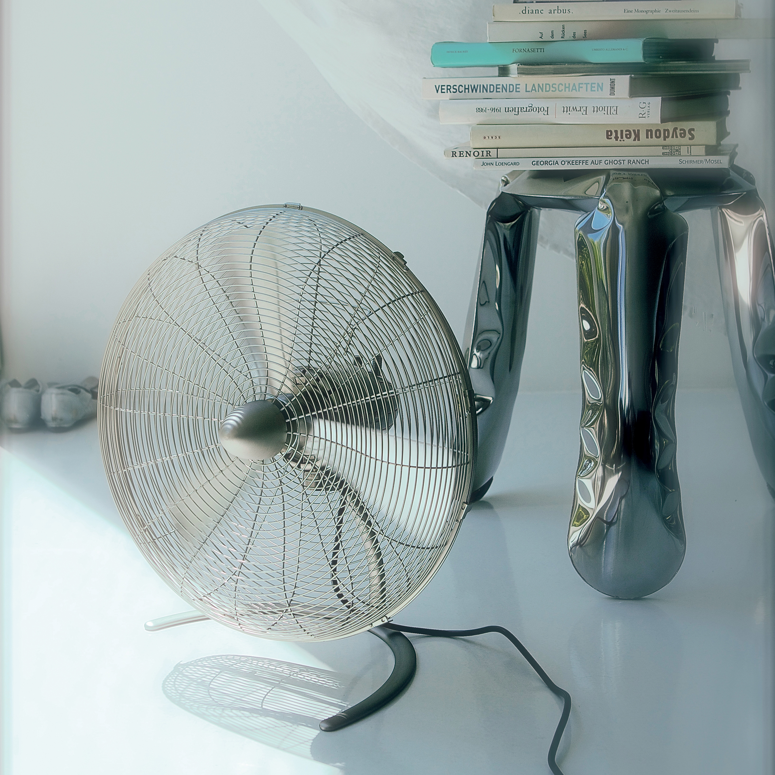 Un ventilateur argenté et un tabouret avec des livres se trouvent près d'une fenêtre avec des rideaux blancs. La lumière est vive.