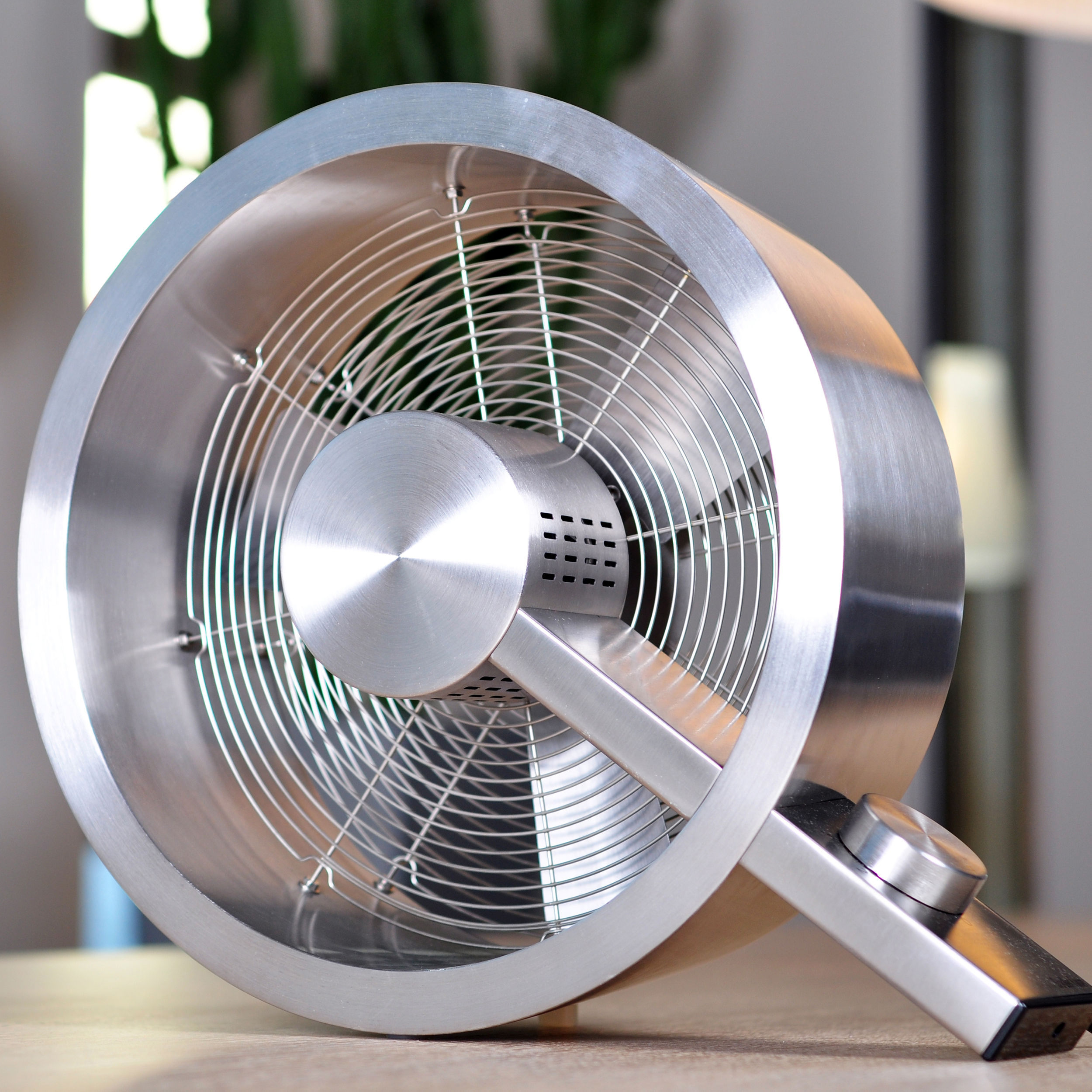 Un ventilateur argenté avec un corps rond et un grillage est posé sur une table en bois, vu sous un angle bas.