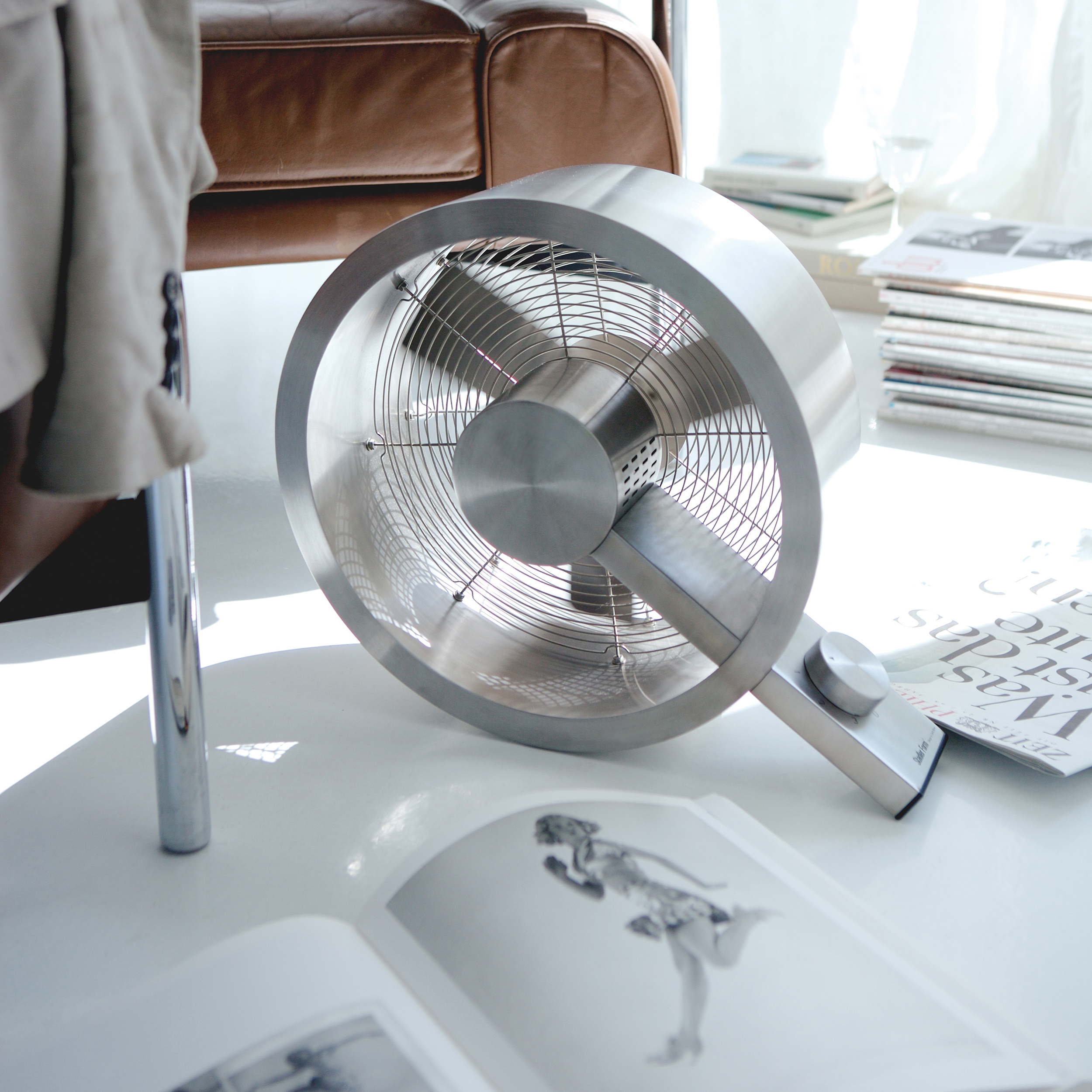 Un petit ventilateur argenté est posé sur une surface blanche près d'une chaise, de livres et de magazines dans une pièce lumineuse.