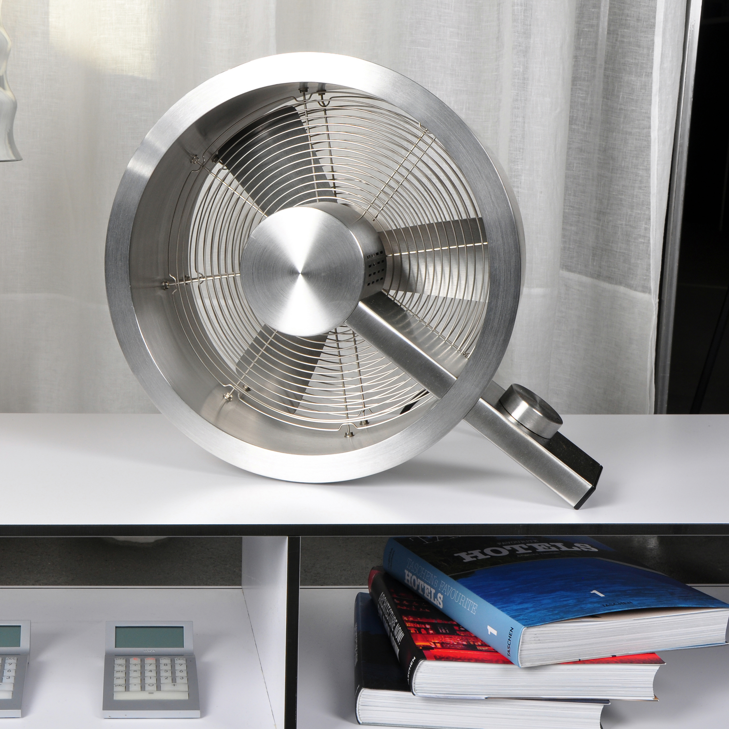 Un ventilateur argenté est posé sur une table blanche avec des livres, des calculatrices et une étagère en arrière-plan.