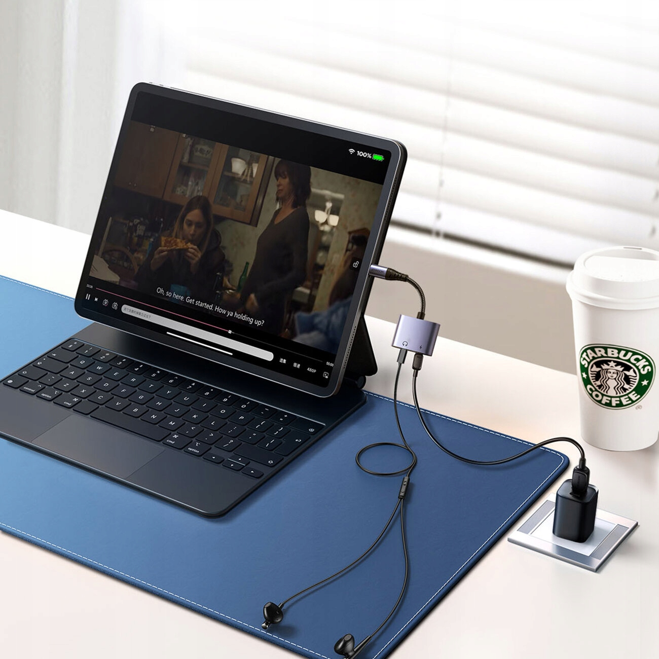 Un tablet con tastiera, adattatore, cuffie e una tazza di Starbucks su un tappetino blu, il tutto su una scrivania bianca.