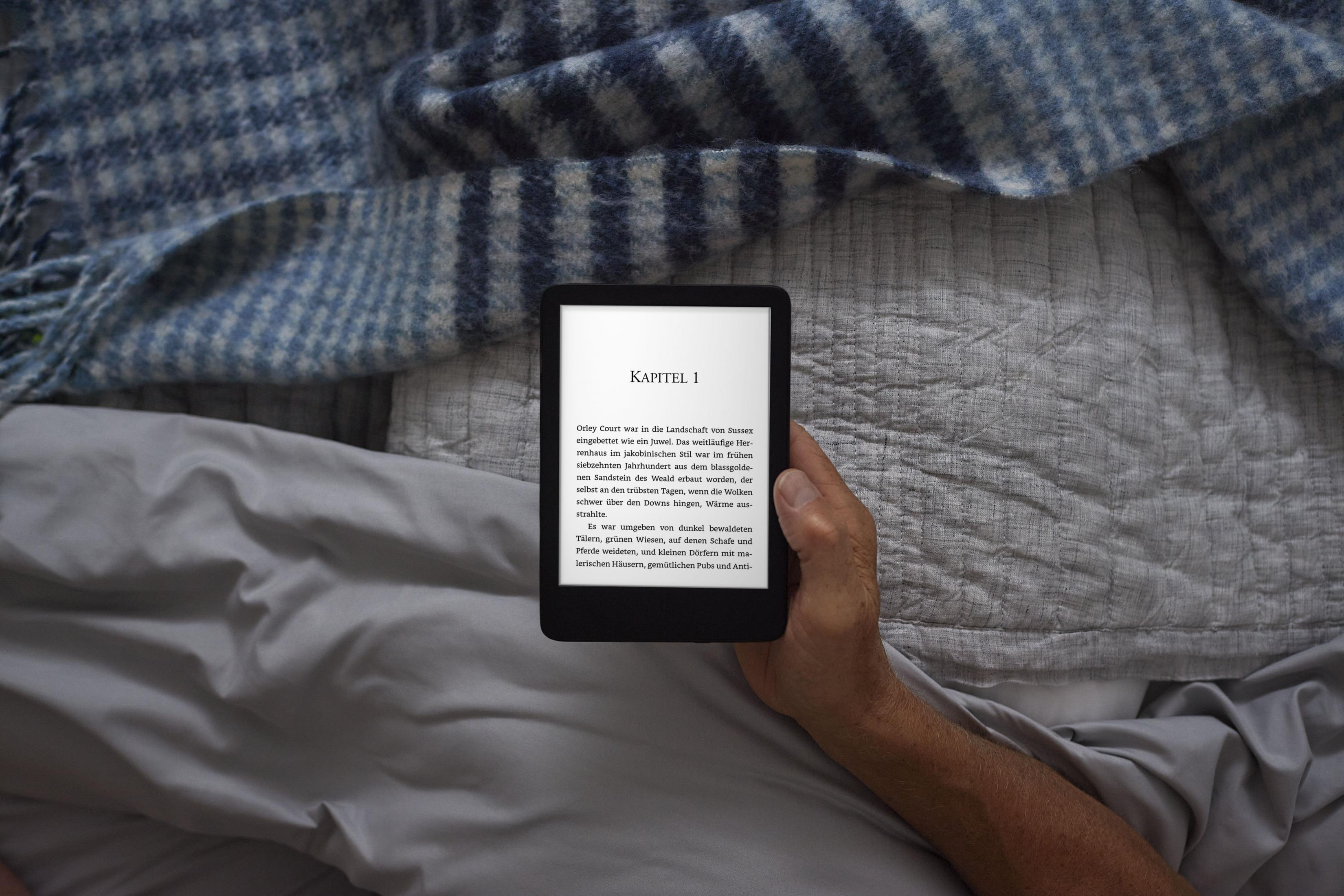 Die Hand einer Person hält einen schwarzen E-Reader im Bett. Der Hintergrund ist ein graues Bettlaken und eine blaue Decke.