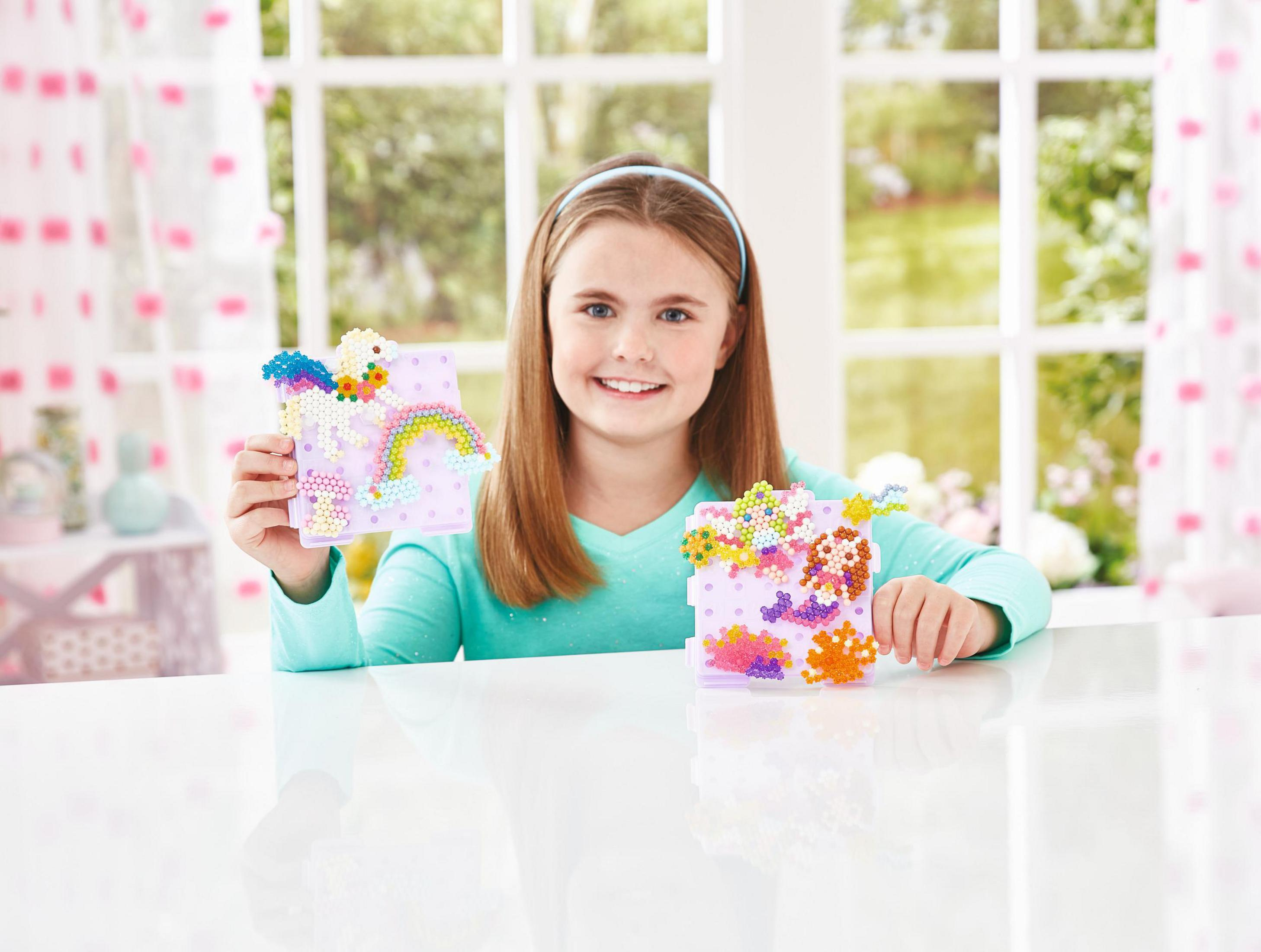 Ein lächelndes Mädchen zeigt Perlenkunst, eine mit einem Einhorn und die andere mit einem farbenfrohen abstrakten Design.