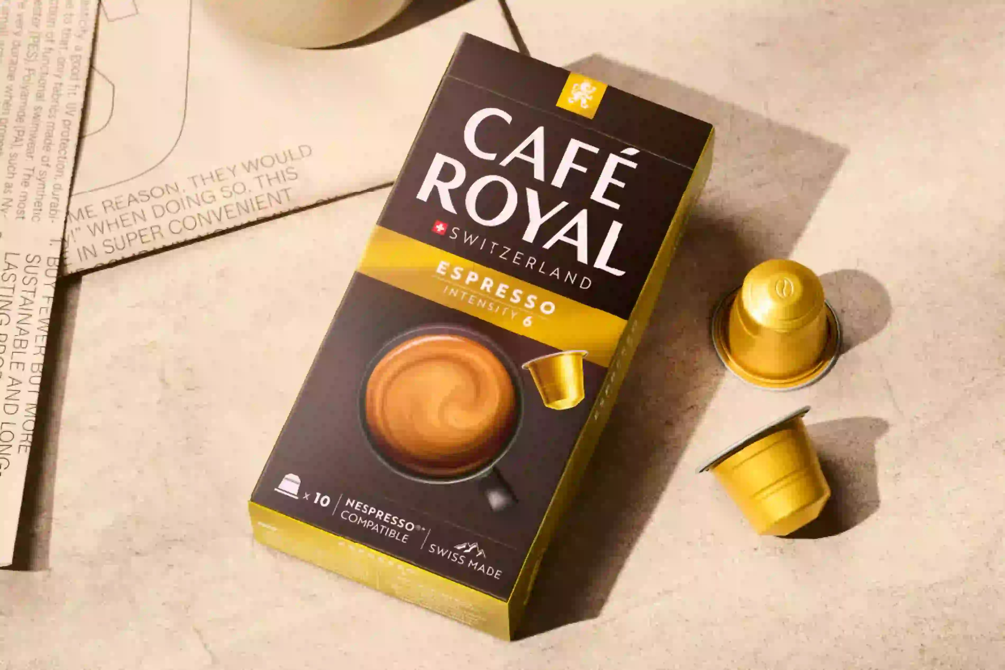 Eine Schachtel Café Royal Espresso-Kapseln mit einem Glas und zwei losen Kapseln auf einer strukturierten Oberfläche.