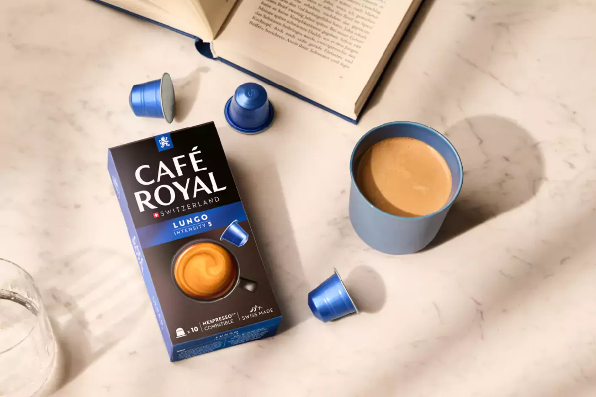 Cafe Royal Lungo-Box, Kaffeetasse und Kapseln auf Marmor. Offenes Buch.