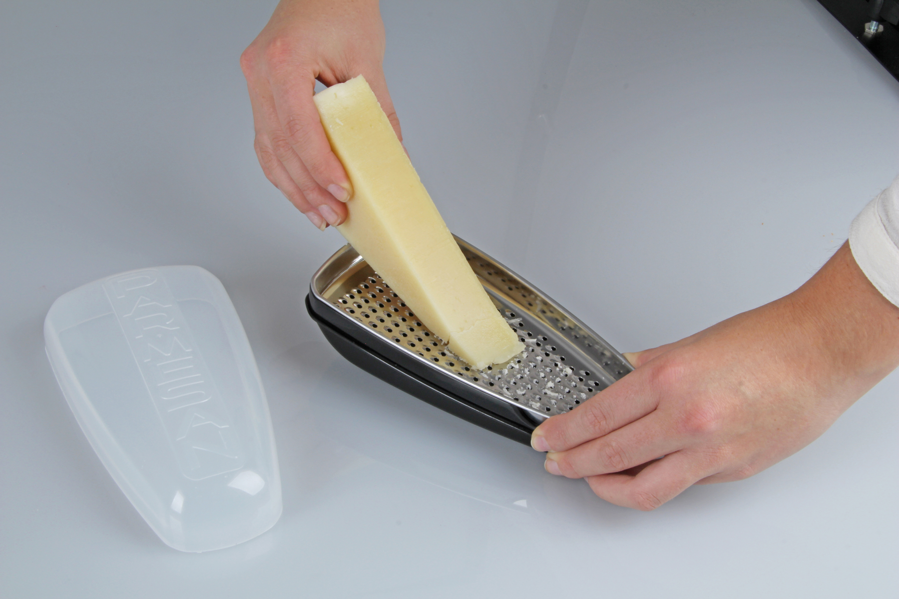 Hände reiben Käse mit einer Reibe mit schwarzem Griff auf einer weißen Oberfläche neben einem transparenten Kunststoffdeckel.