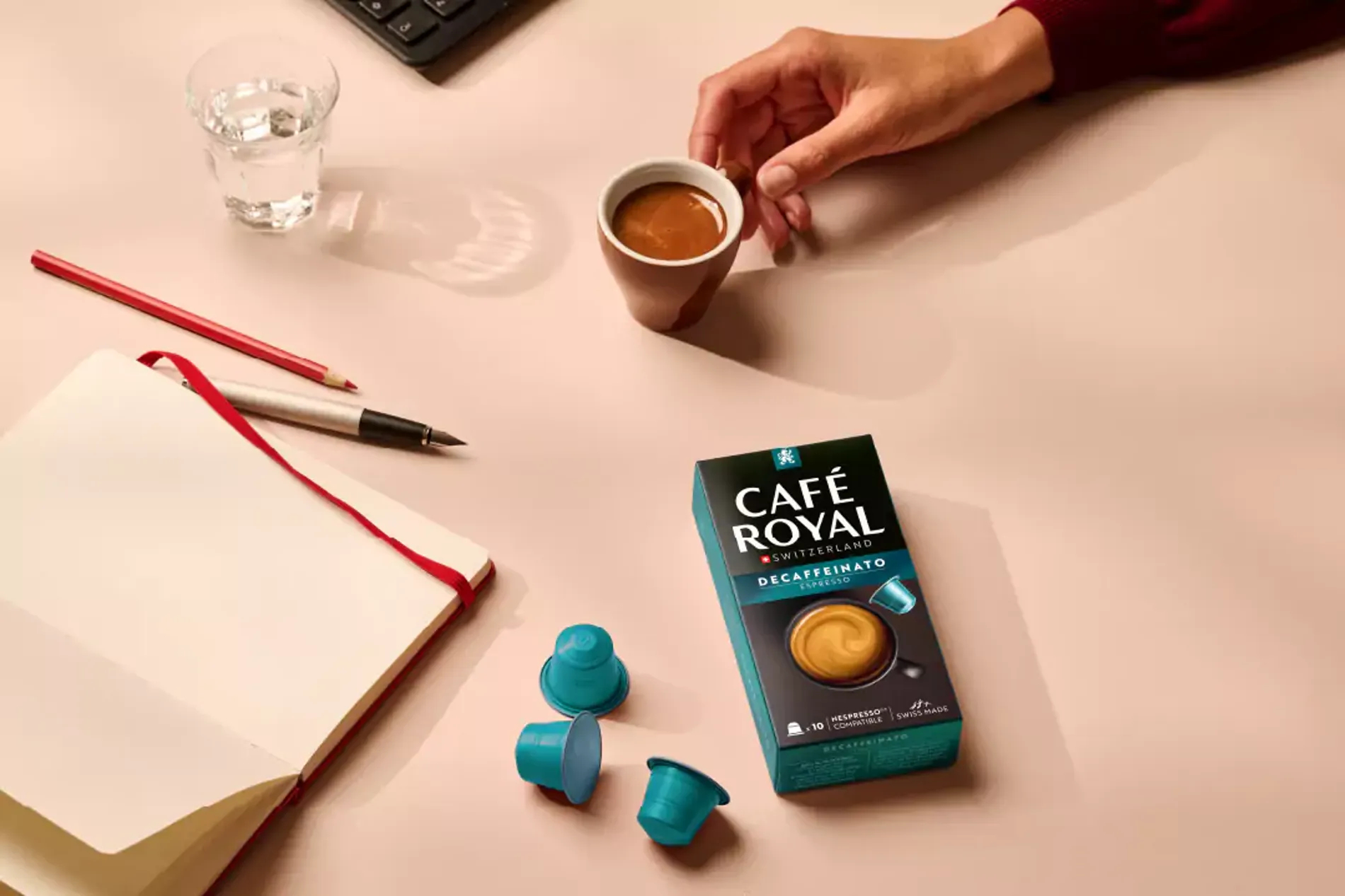 Eine Schreibtischszene zeigt eine Café Royal Kaffeebox, Kapseln, Notizbuch, Stift, ein Glas Wasser und eine Hand, die eine Kaffeetasse hält.