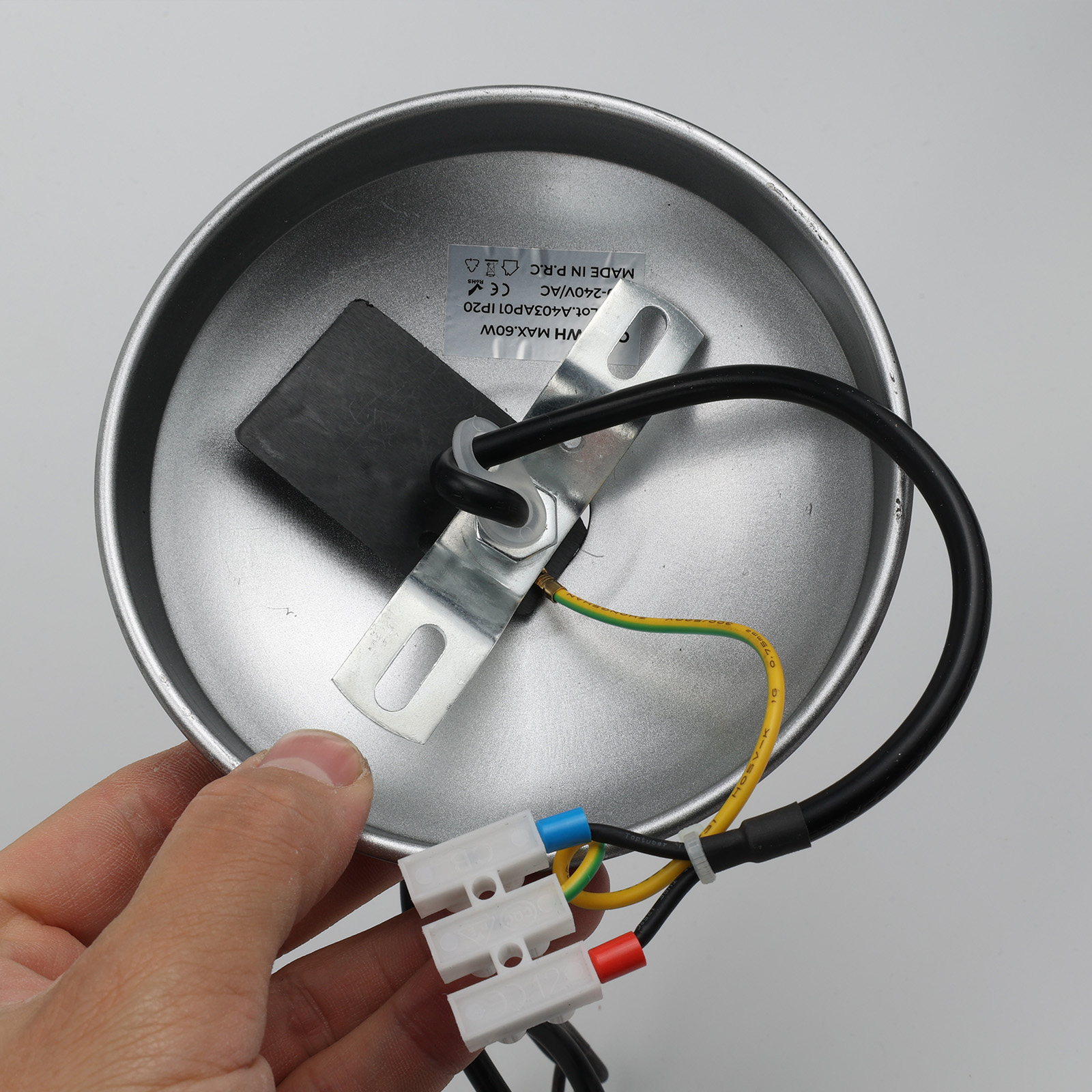Una mano sostiene la parte superior de una lámpara colgante. Se ven cables y conectores. El fondo es gris claro.