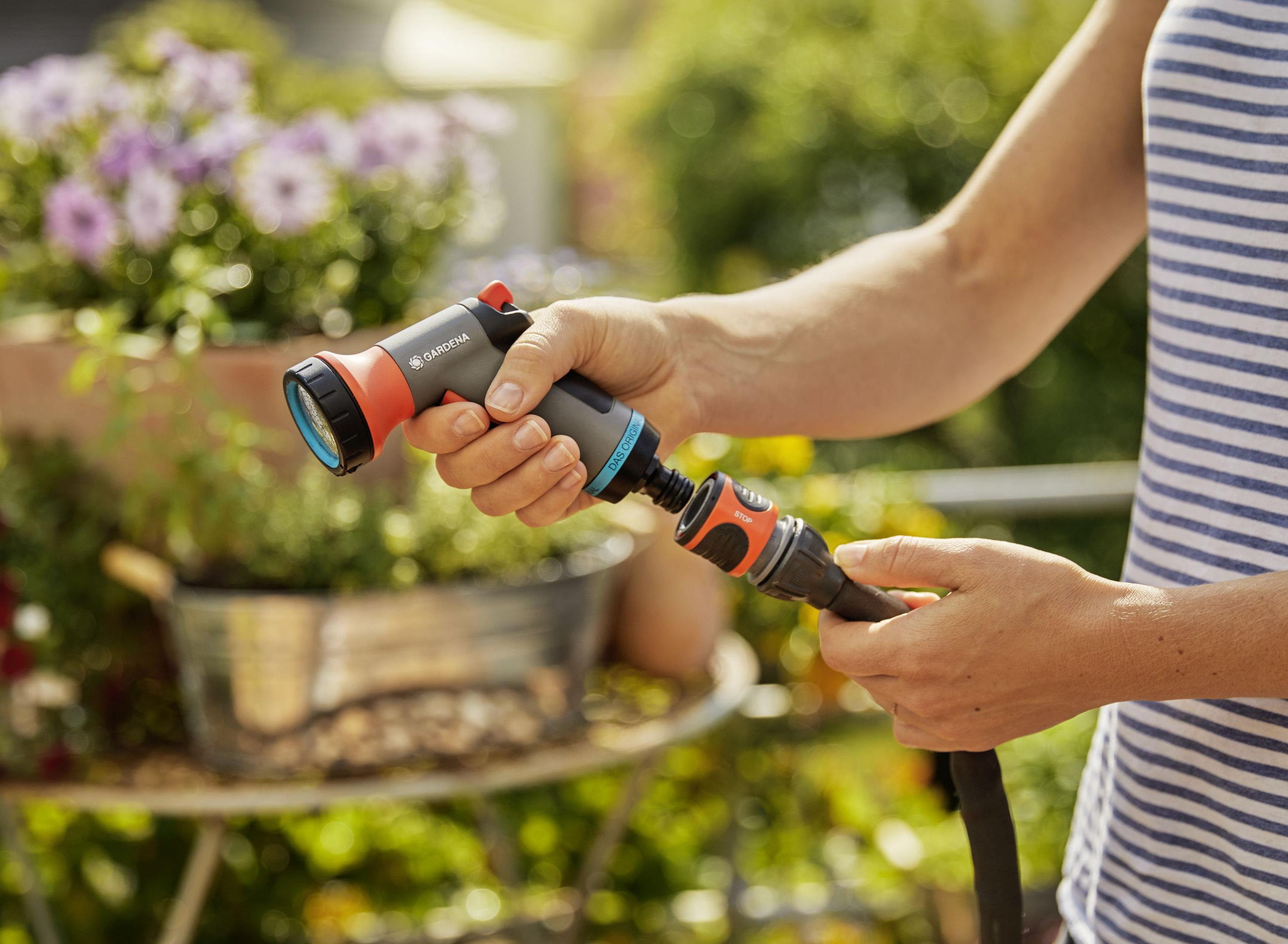 Eine Person verbindet eine Gardena Sprühdüse mit einem Schlauch, bereit, Pflanzen in einer Gartenumgebung zu bewässern.