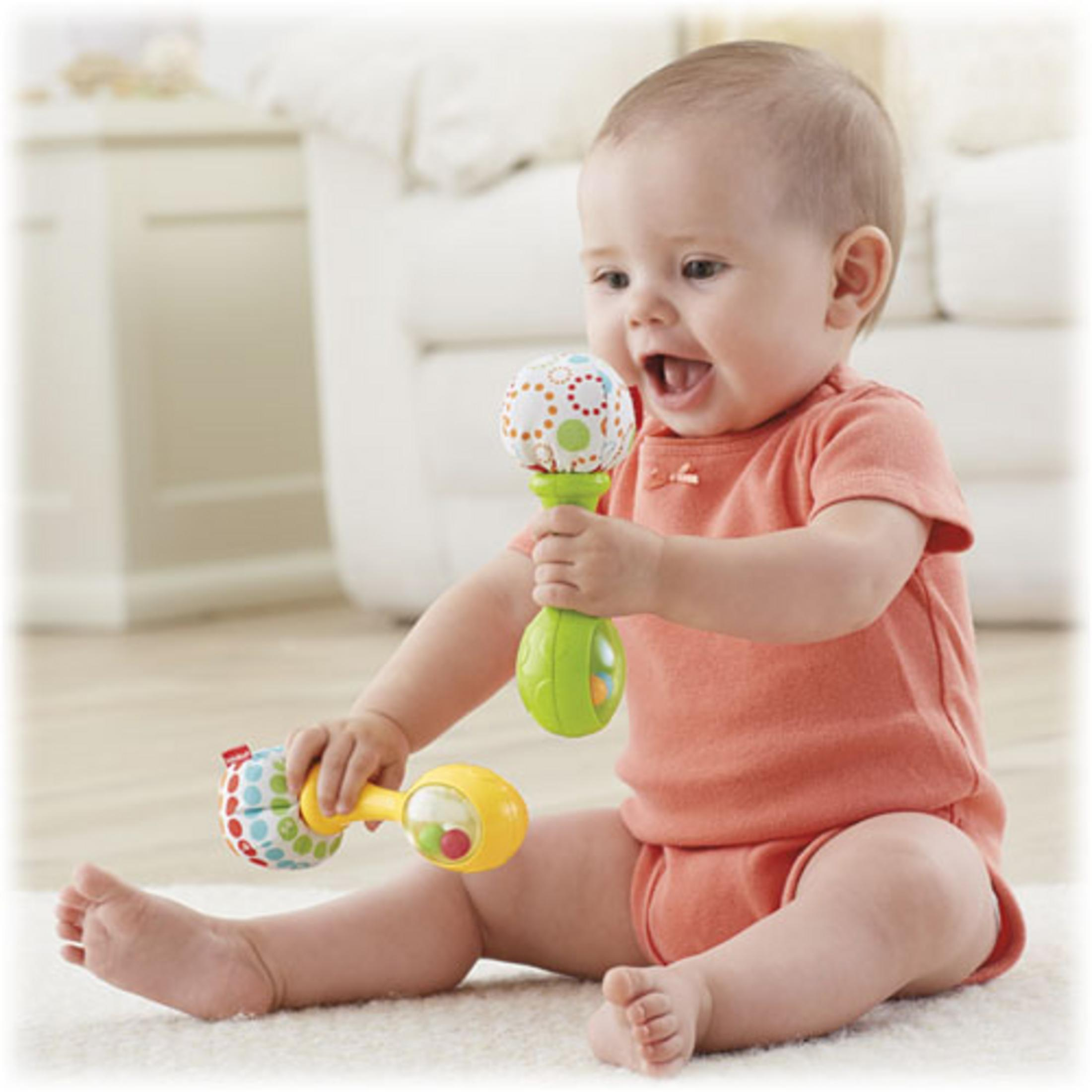Baby in pfirsichfarbenem Hemd hält grüne und gelbe Maracas und lächelt breit.