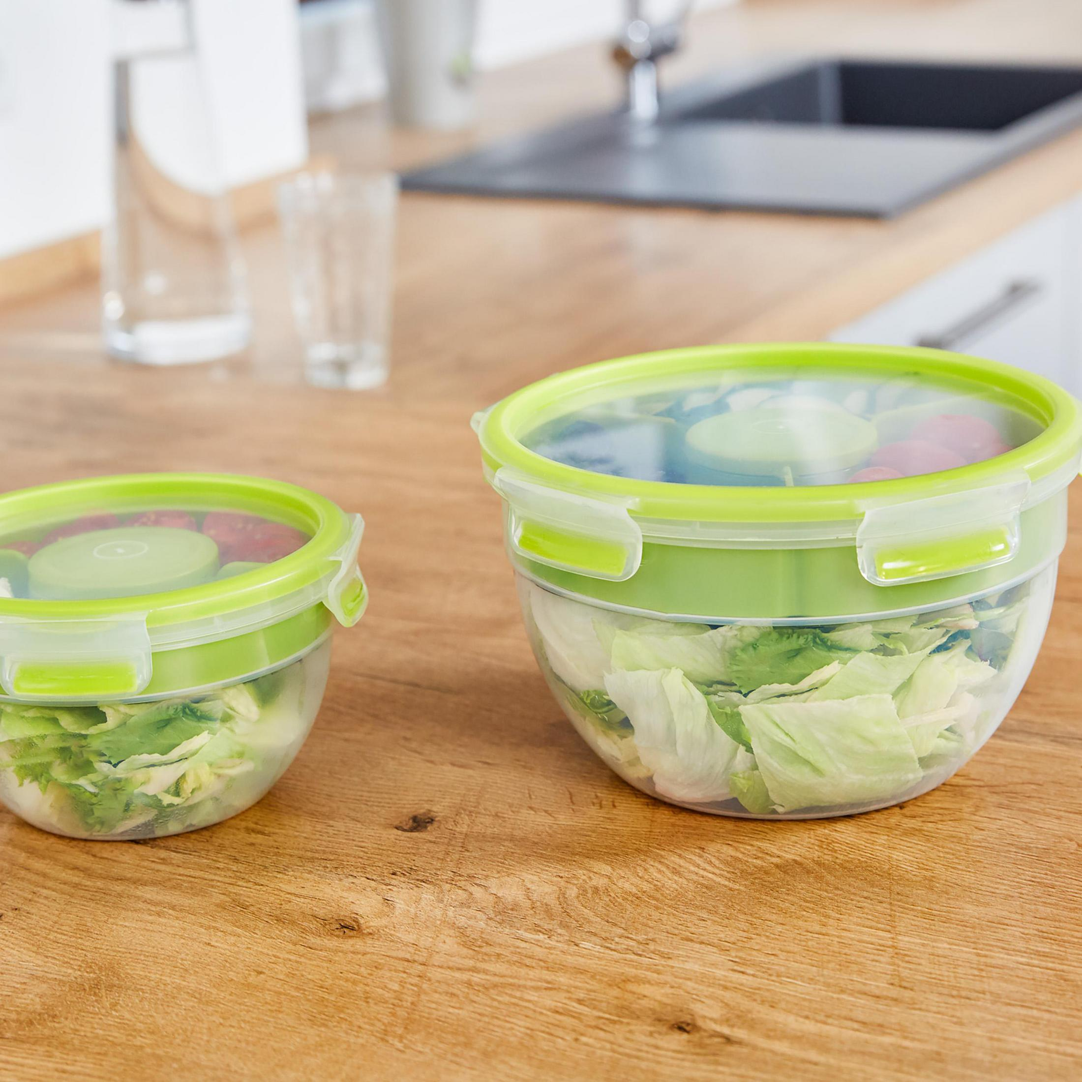 Zwei Schalen mit Salat. Sie haben grüne Deckel. Sie stehen auf einer Holzoberfläche, neben einem Waschbecken und einer Vase.