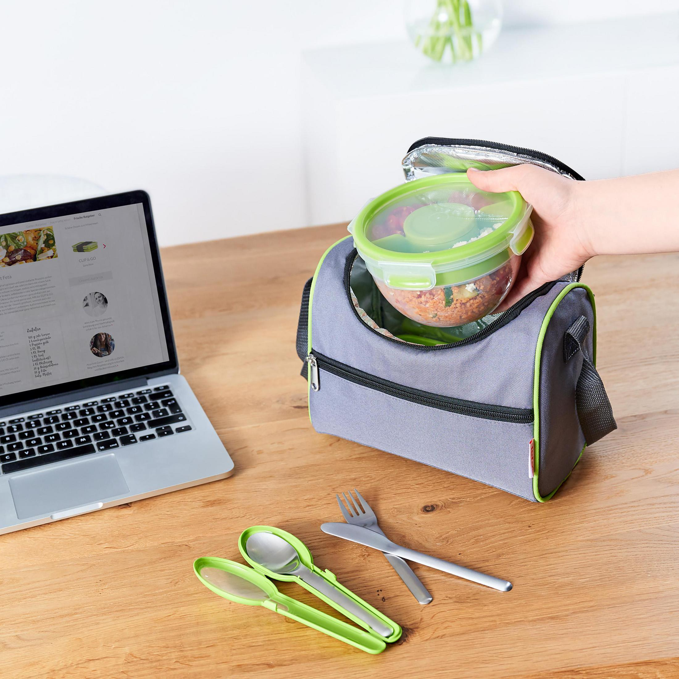 Eine Person legt einen Lebensmittelbehälter in eine graue Lunchtasche. Auf dem Tisch liegen Utensilien und ein Laptop.