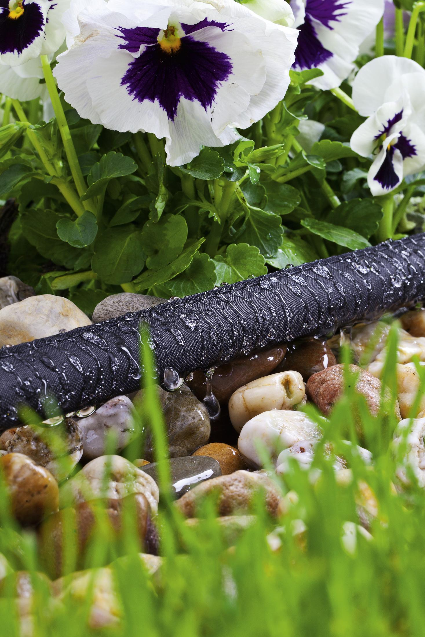 Schwarzer Schlauch mit Wassertröpfchen, umgeben von Kieselsteinen und Blumen.