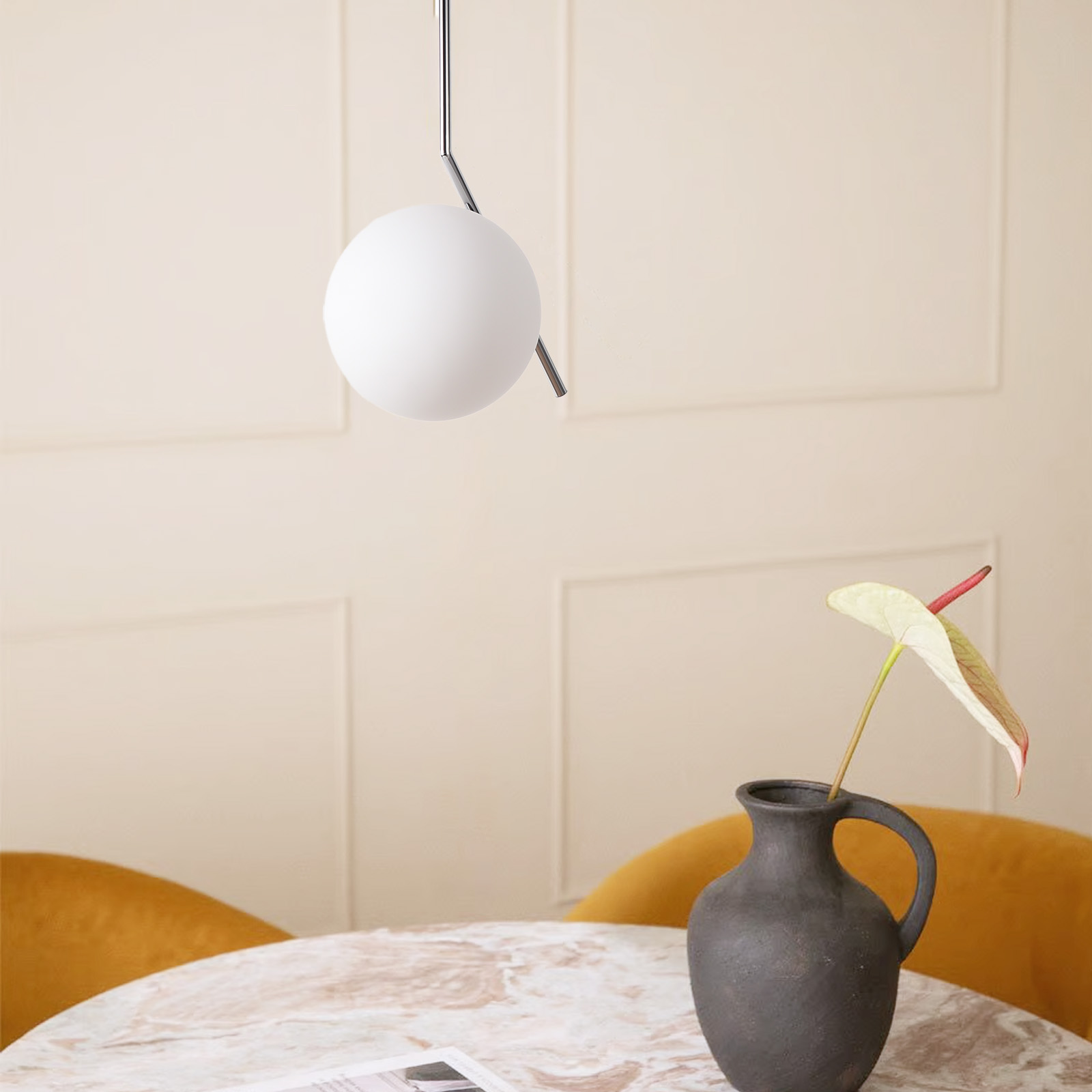 Una lámpara colgante blanca cuelga sobre una mesa de mármol con un jarrón que sostiene una flor, contra una pared de color amarillo claro.