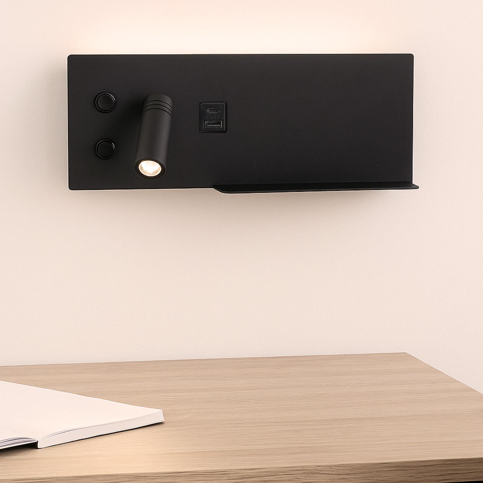 Una lámpara de pared negra con un foco, puertos USB y un pequeño estante montado sobre un escritorio de madera con un cuaderno.