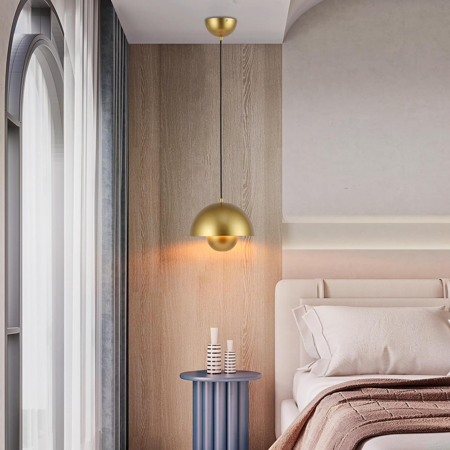 Una lámpara colgante dorada cuelga sobre una mesa azul junto a una cama en una habitación con luz natural.