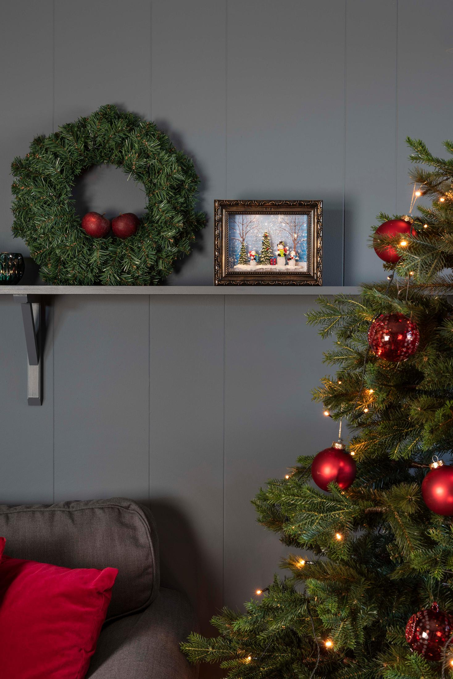 Weihnachtsdeko: Kranz, Schneekugelbild, Baum mit Ornamenten, Sofa mit roten Kissen vor grauer Wand.