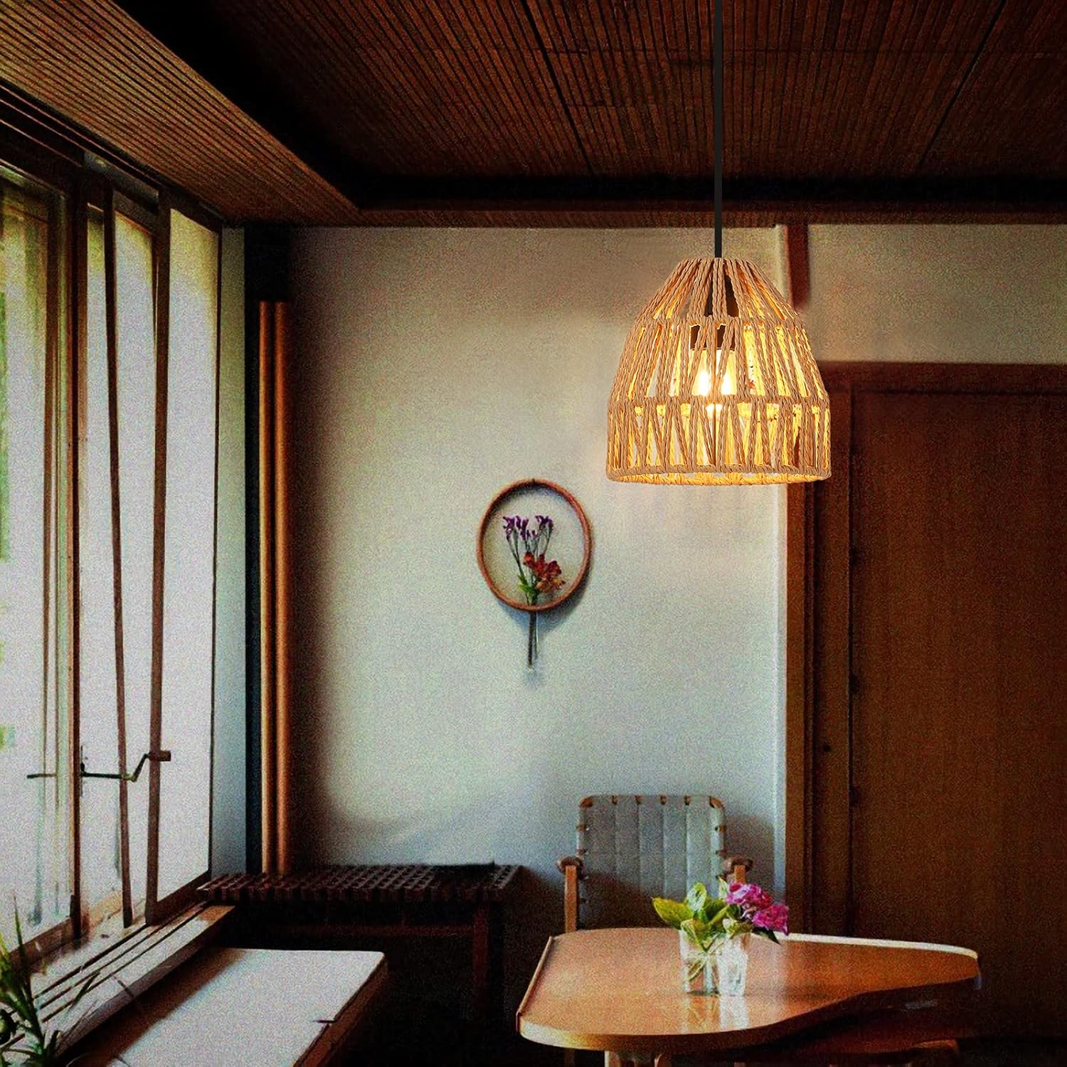 Una lámpara tejida cuelga en una habitación con mesa, silla, flores y ventana, creando un espacio acogedor.