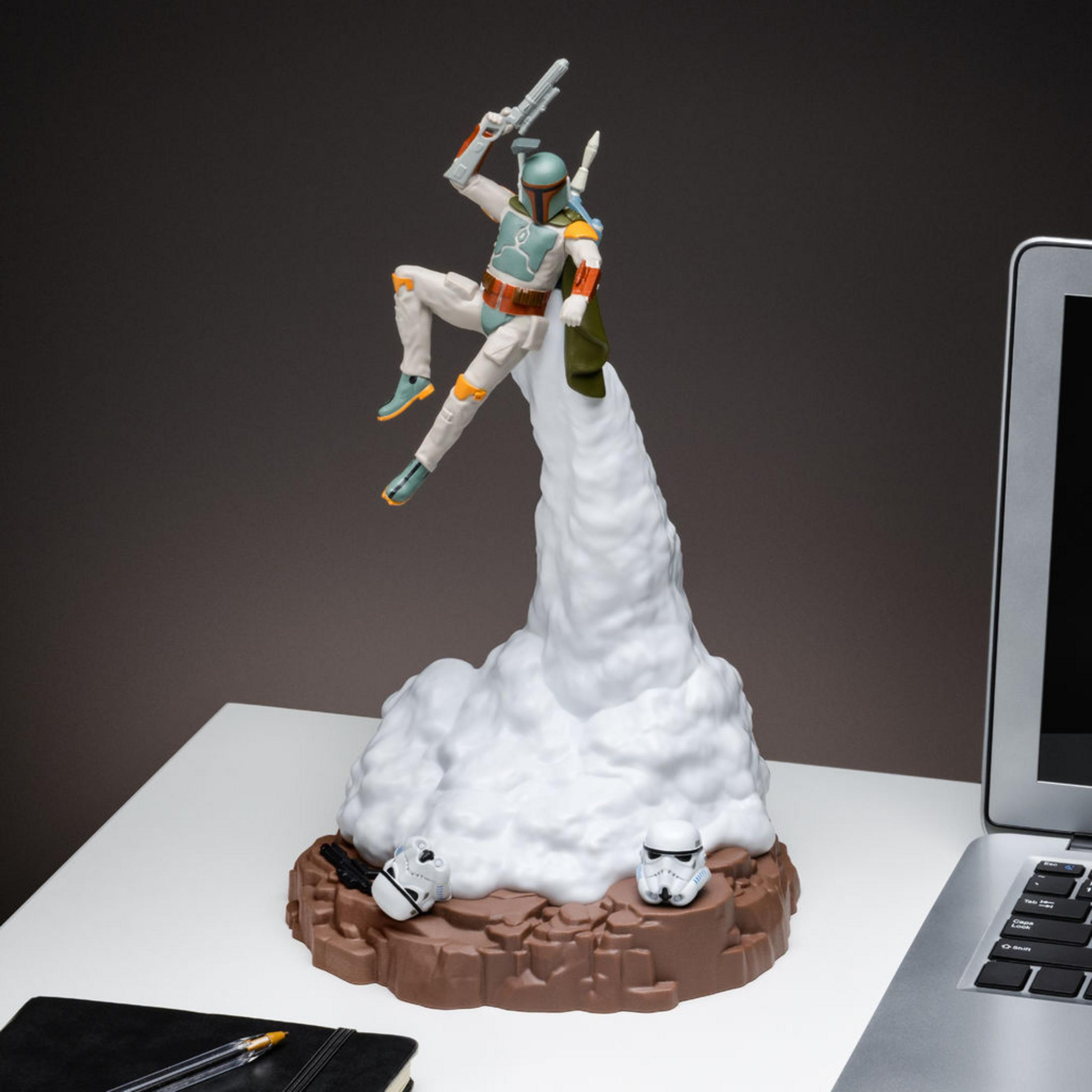 Boba Fett Figur springt von Rauchwolke, flankiert von Sturmtruppenhelmen, auf einem Schreibtisch in der Nähe eines Laptops.