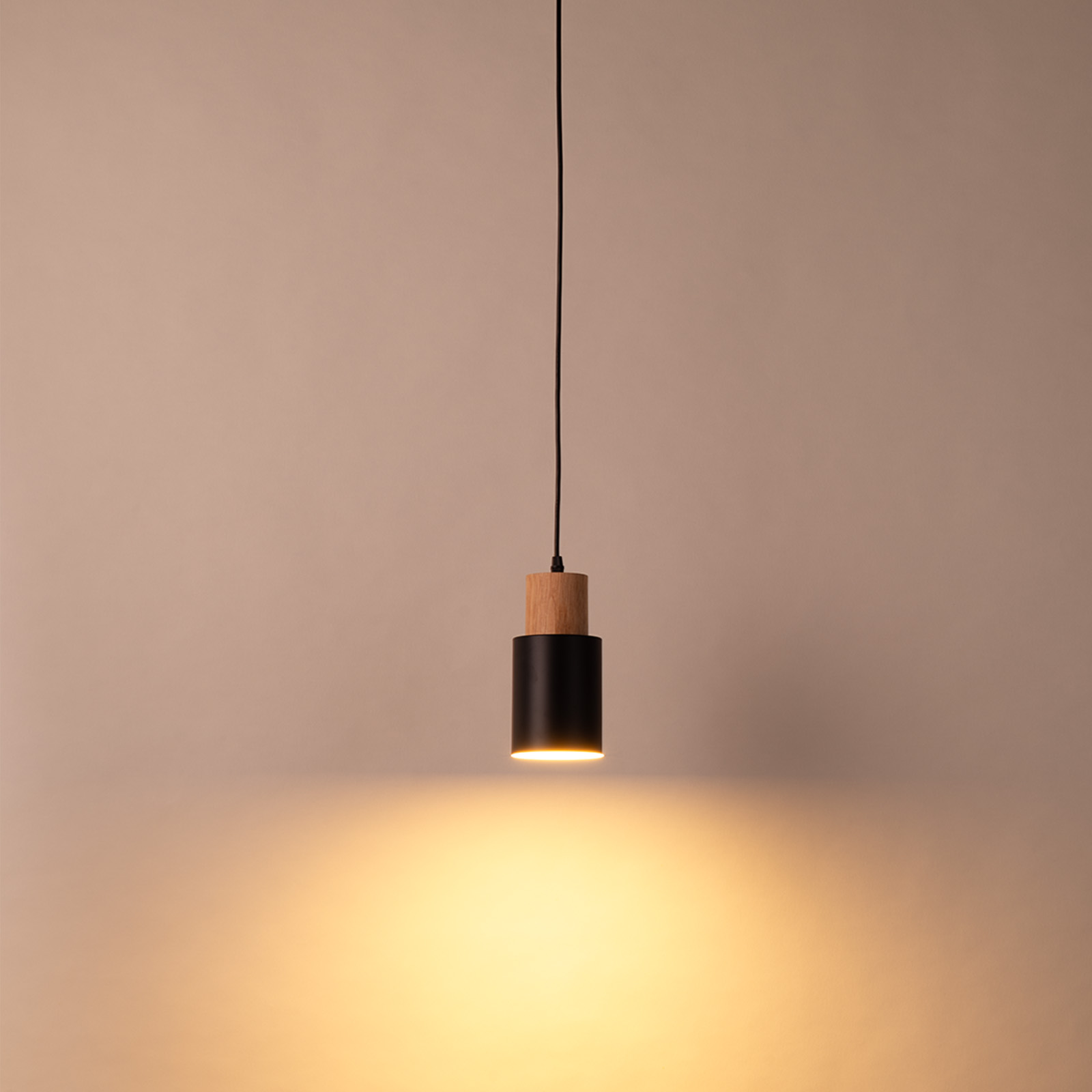 Una lámpara colgante negra y de madera cuelga, iluminada desde abajo, sobre una pared neutra.