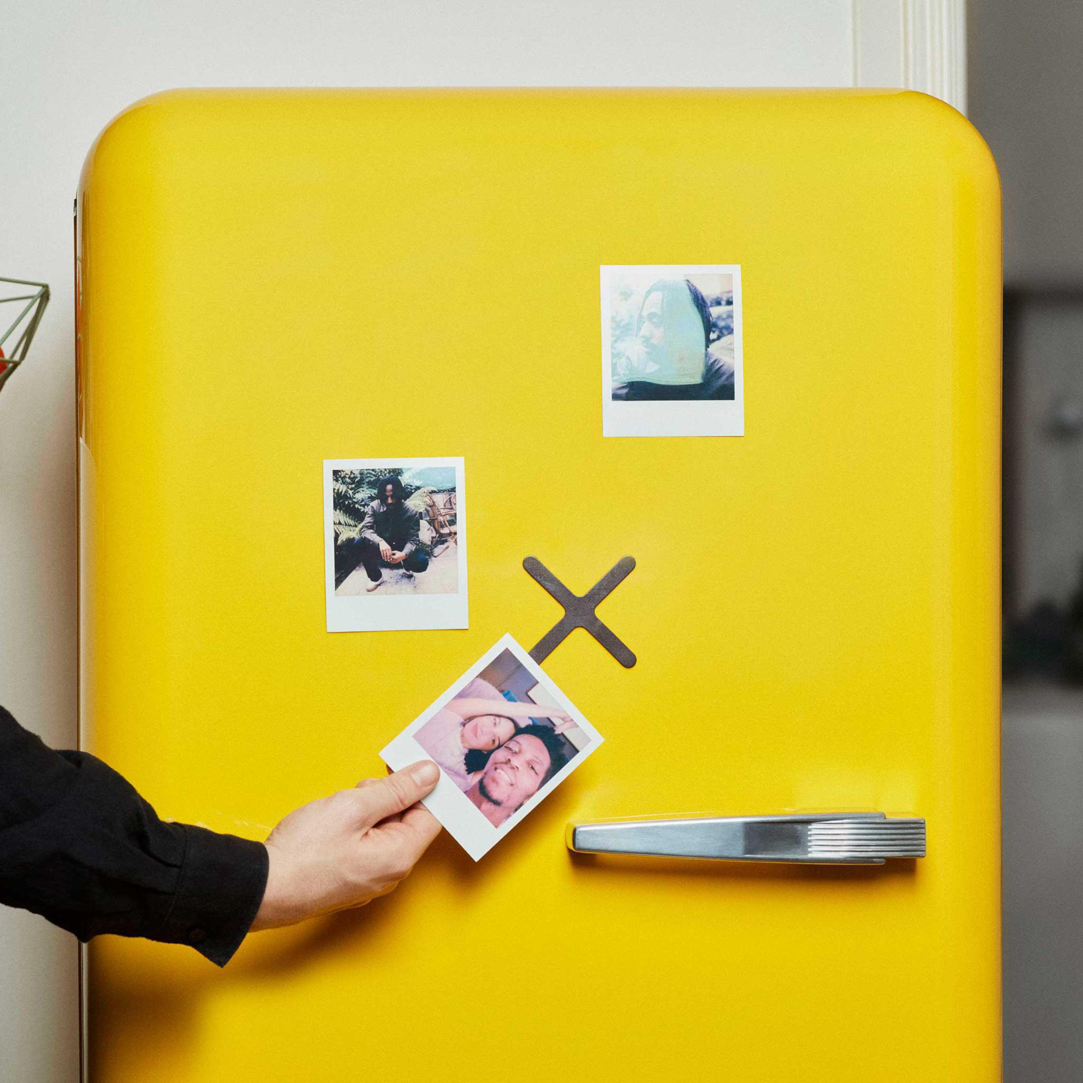 Un frigorifero giallo con foto istantanee allegate, una delle quali è tenuta in mano da una mano in una manica nera.