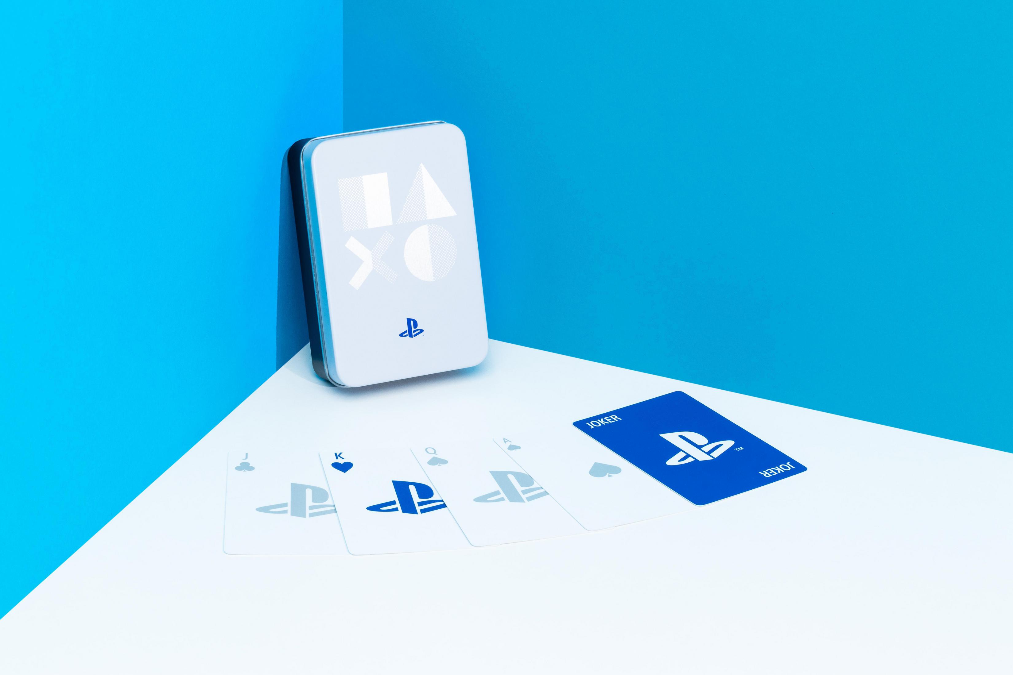 Auf einer weißen Oberfläche vor einer blauen Ecke sind Spielkarten im PlayStation-Design und ein Kartenetui angeordnet.