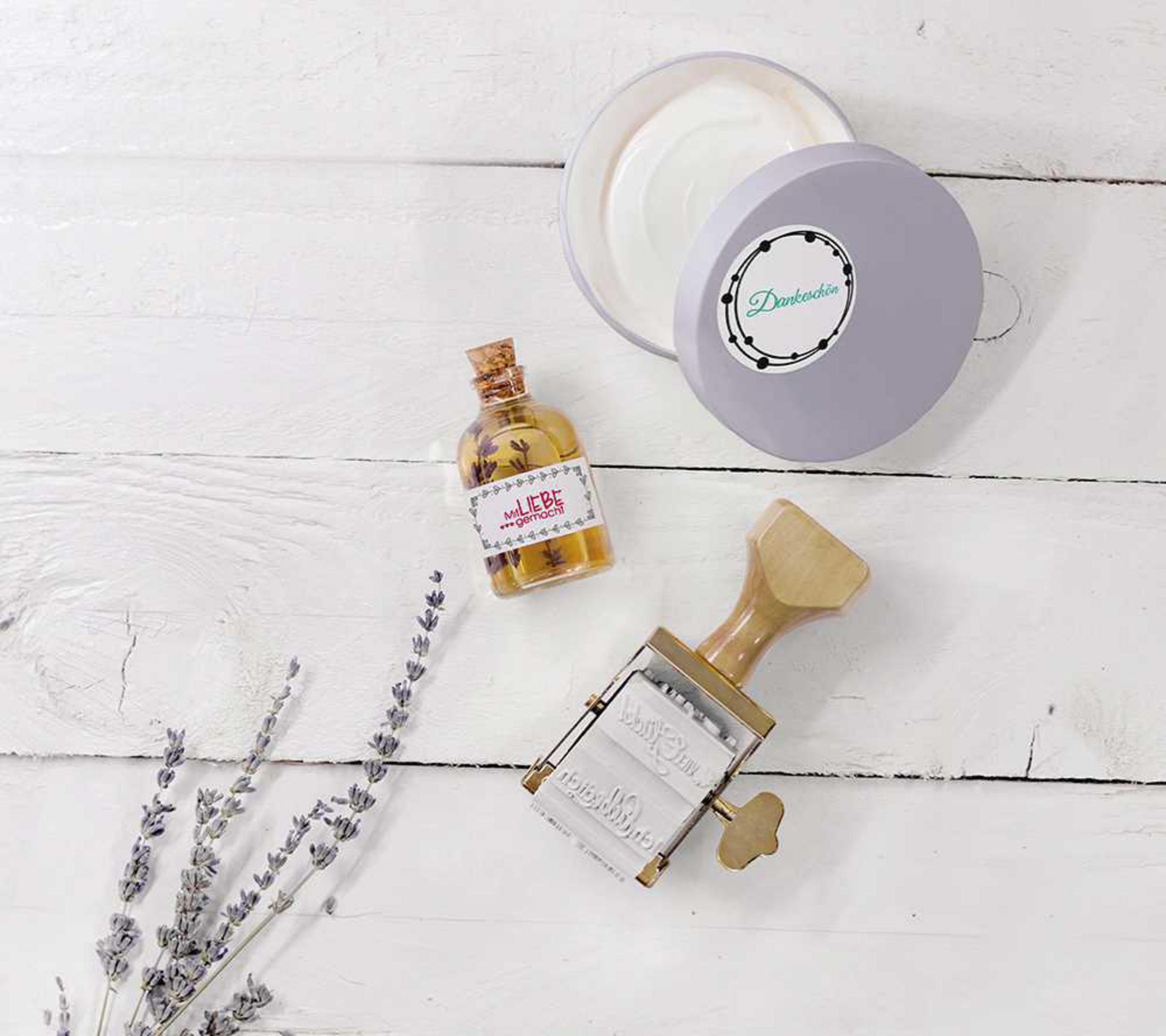 Ein Stempel, Lavendel, eine Ölflasche und eine Creme sitzen auf weißem Holz. Der Stempel hat einen Holzgriff.