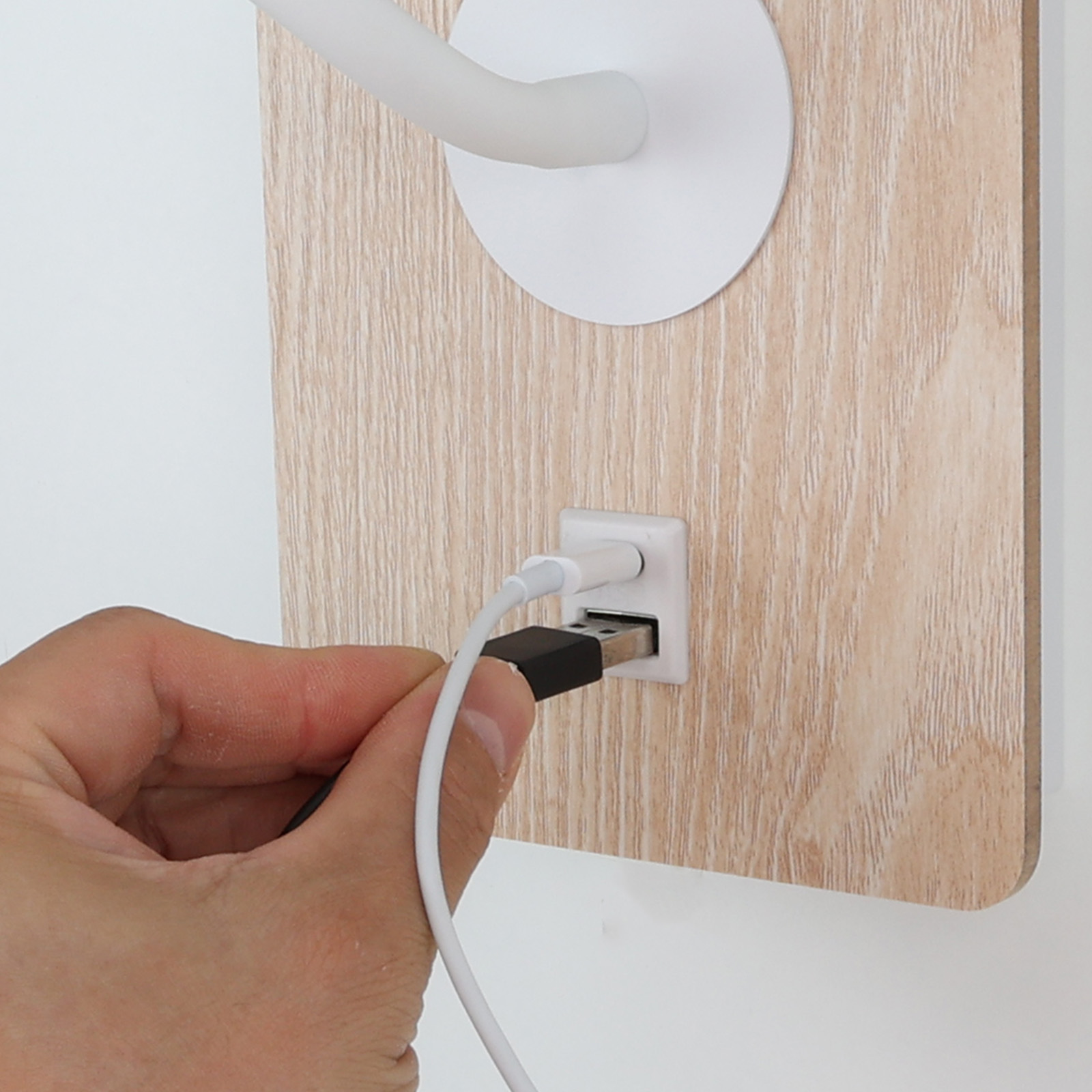 Una mano conecta un cable USB blanco a un soporte de pared de madera con una lámpara blanca. El fondo es una pared blanca.