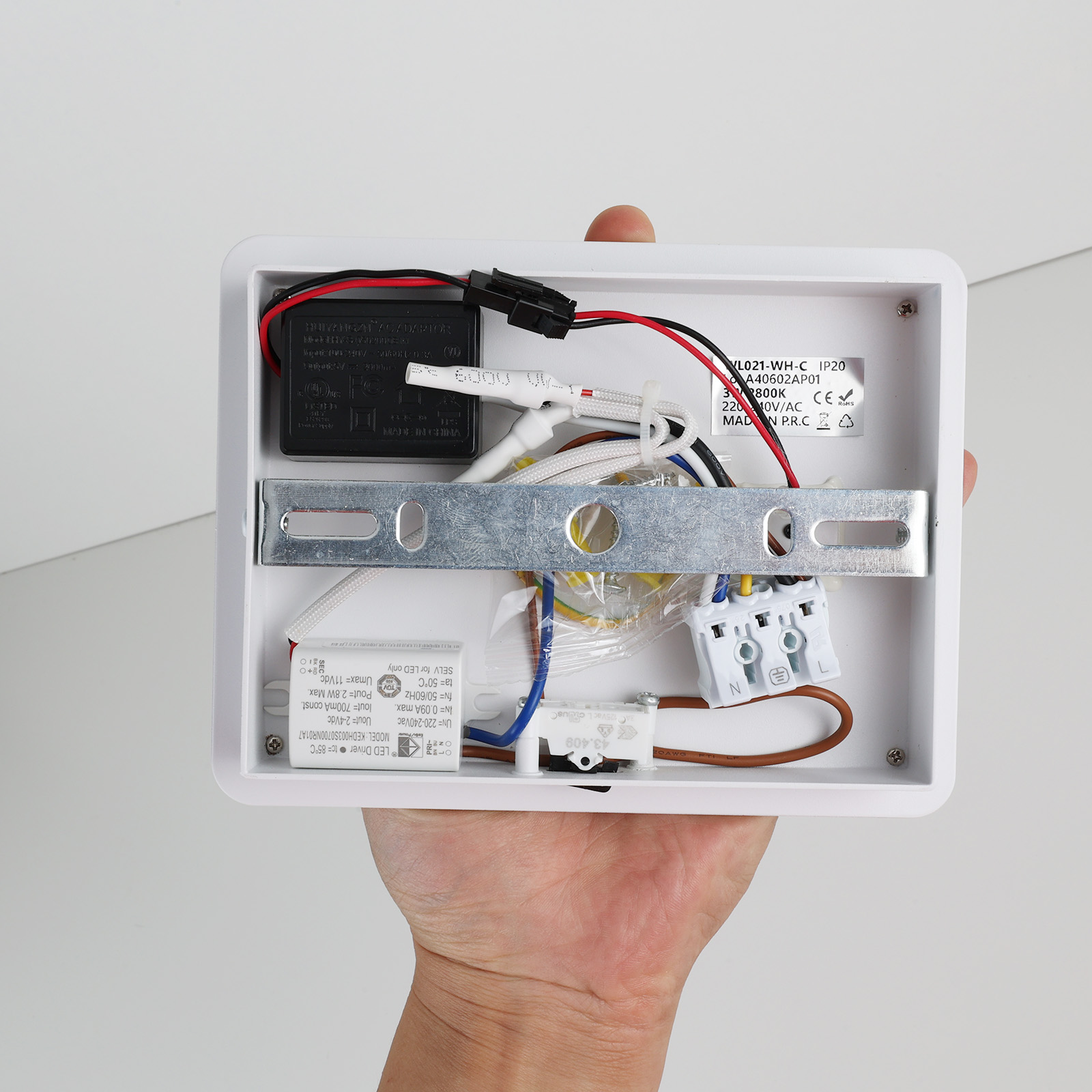 Una mano sostiene una caja eléctrica blanca abierta, revelando cableado, circuitos y etiquetas en el interior, sobre un fondo blanco.