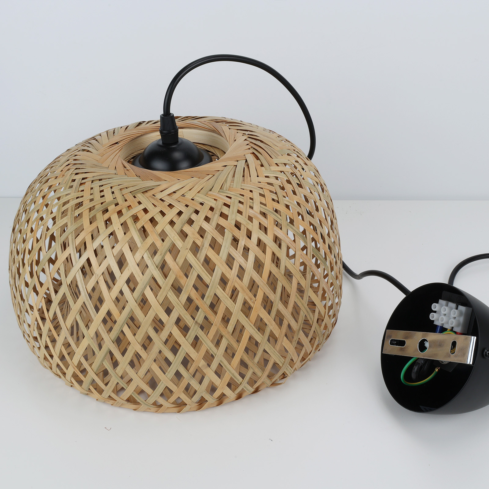 Lámpara colgante de bambú tejido con herrajes y cableado negros sobre una superficie blanca.