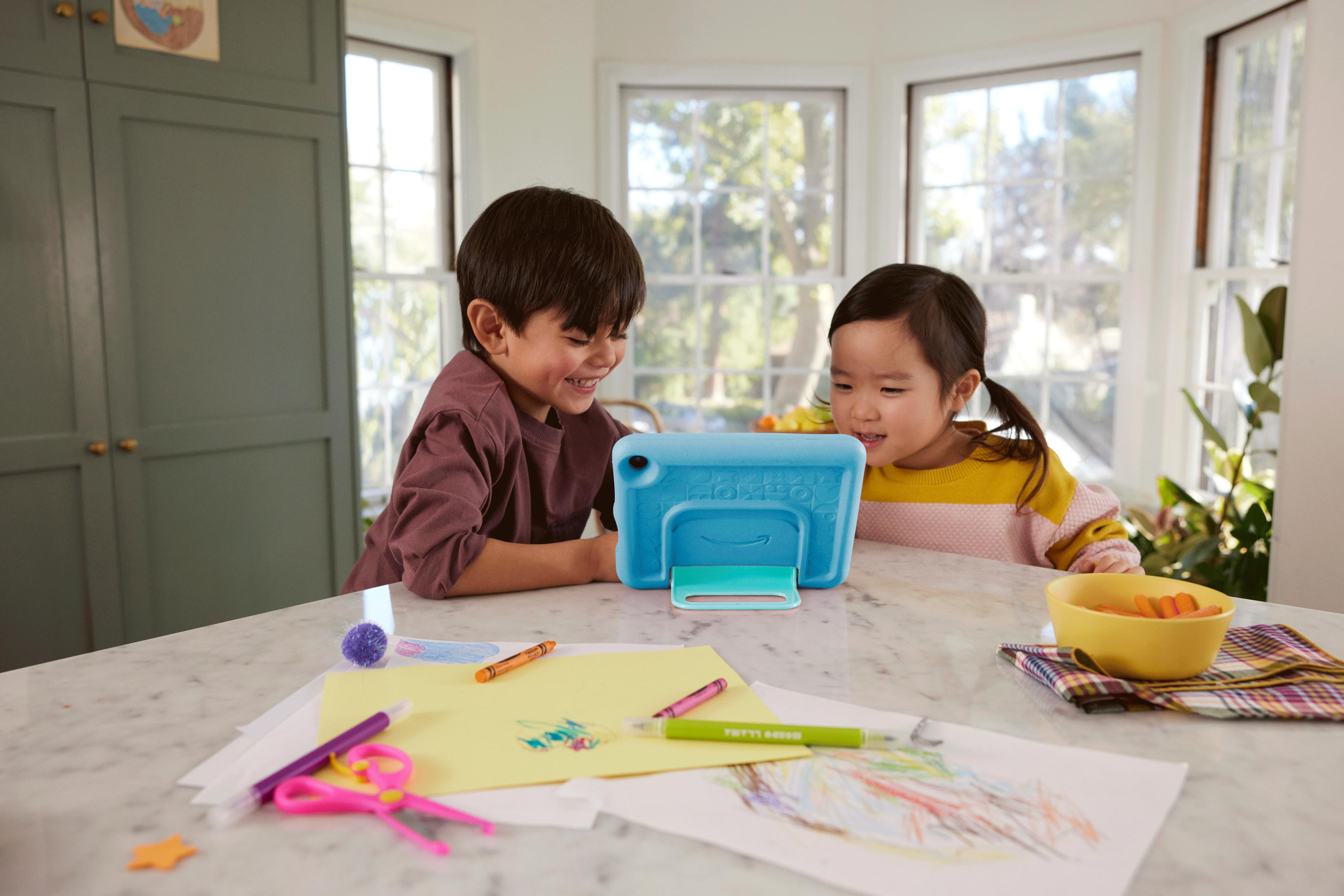 Due bambini che guardano un tablet blu su un tavolo di marmo con materiale artistico sparso intorno.