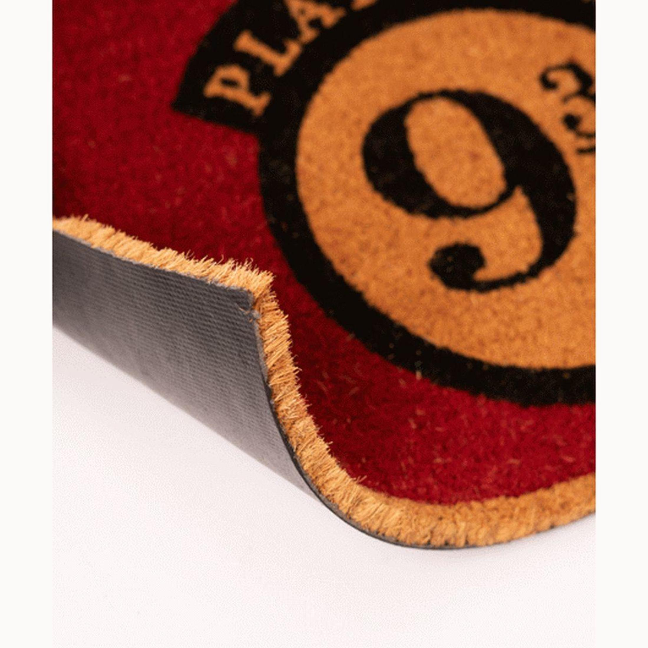 Nahaufnahme einer rot-braunen 'Platform 9 3/4'-Fußmatte, die Textur und Rückseite zeigt. Ecke ist angehoben.