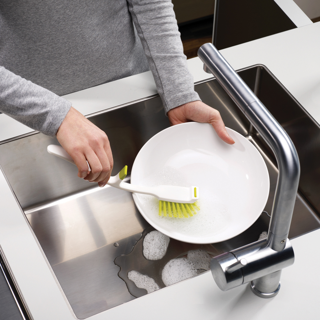 Eine Person wäscht einen weißen Teller mit einer grün-weißen Bürste in einem Edelstahlspülbecken mit einem silbernen Wasserhahn.