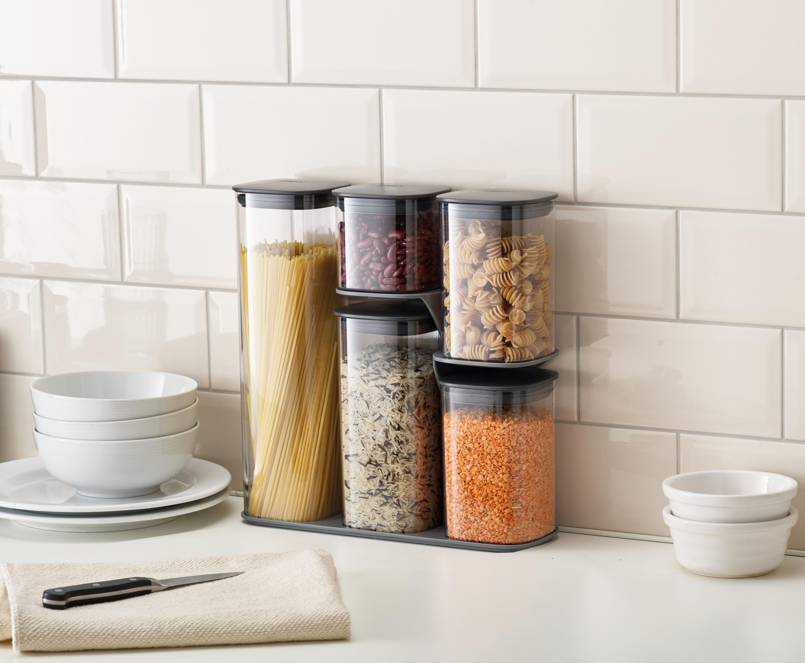 Recipientes de almacenamiento transparentes con pasta, frijoles y lentejas, dispuestos contra un protector contra salpicaduras.